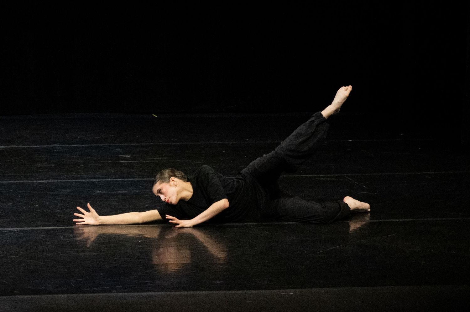 A dancer in black attire performs a graceful floor movement on a dark stage with arms outstretched.