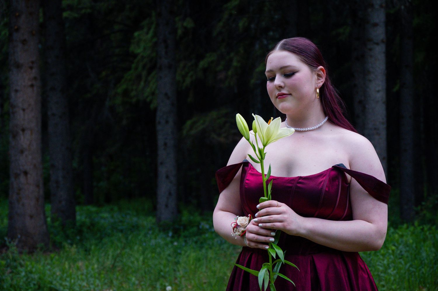 Someone in a burgundy off-shoulder dress holds white lilies while standing in front of a forest backdrop at dusk.