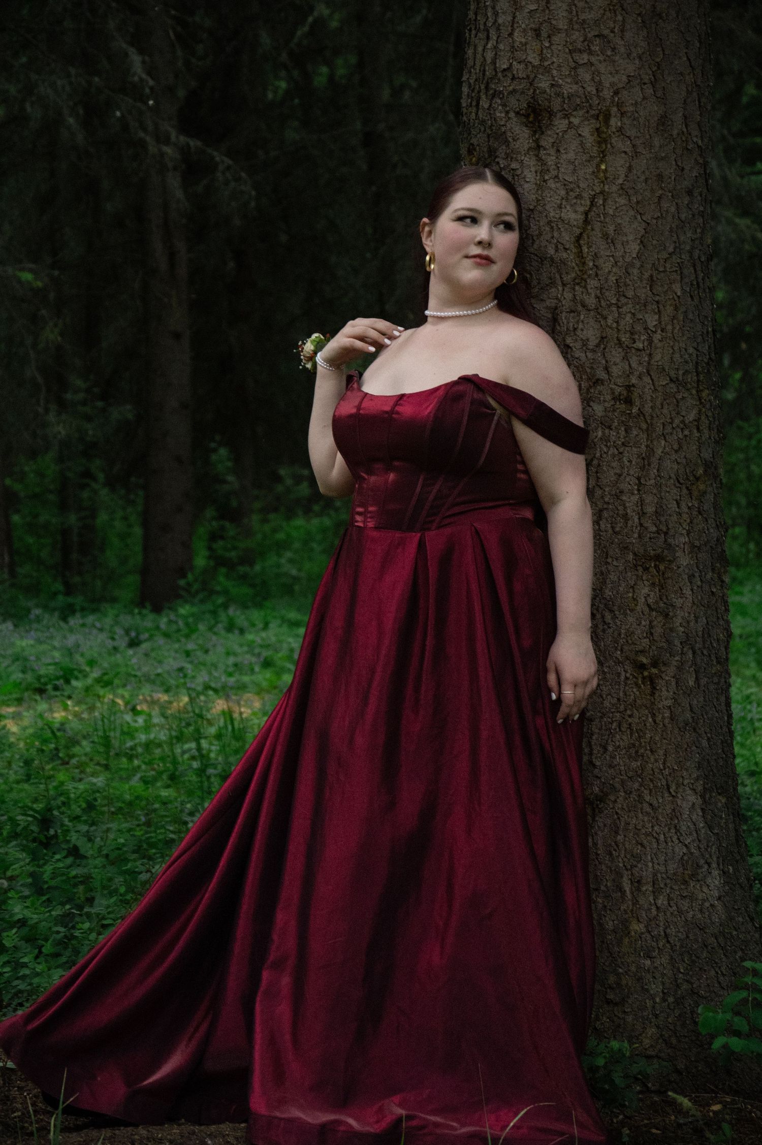 A person in a flowing burgundy velvet gown poses gracefully against a tree trunk in a serene forest setting.