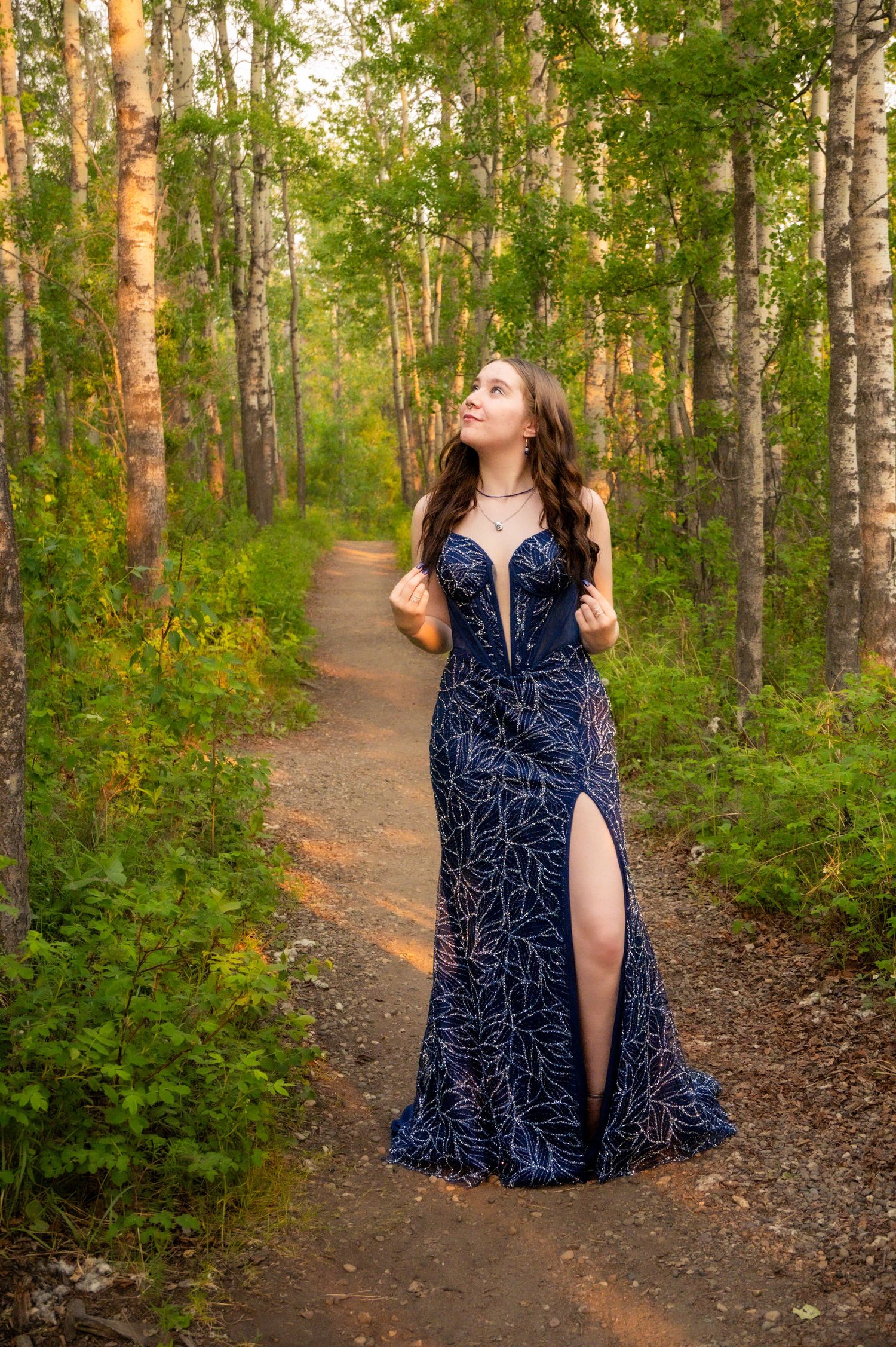 A person in a navy patterned dress poses along a sunlit forest path lined with birch trees.