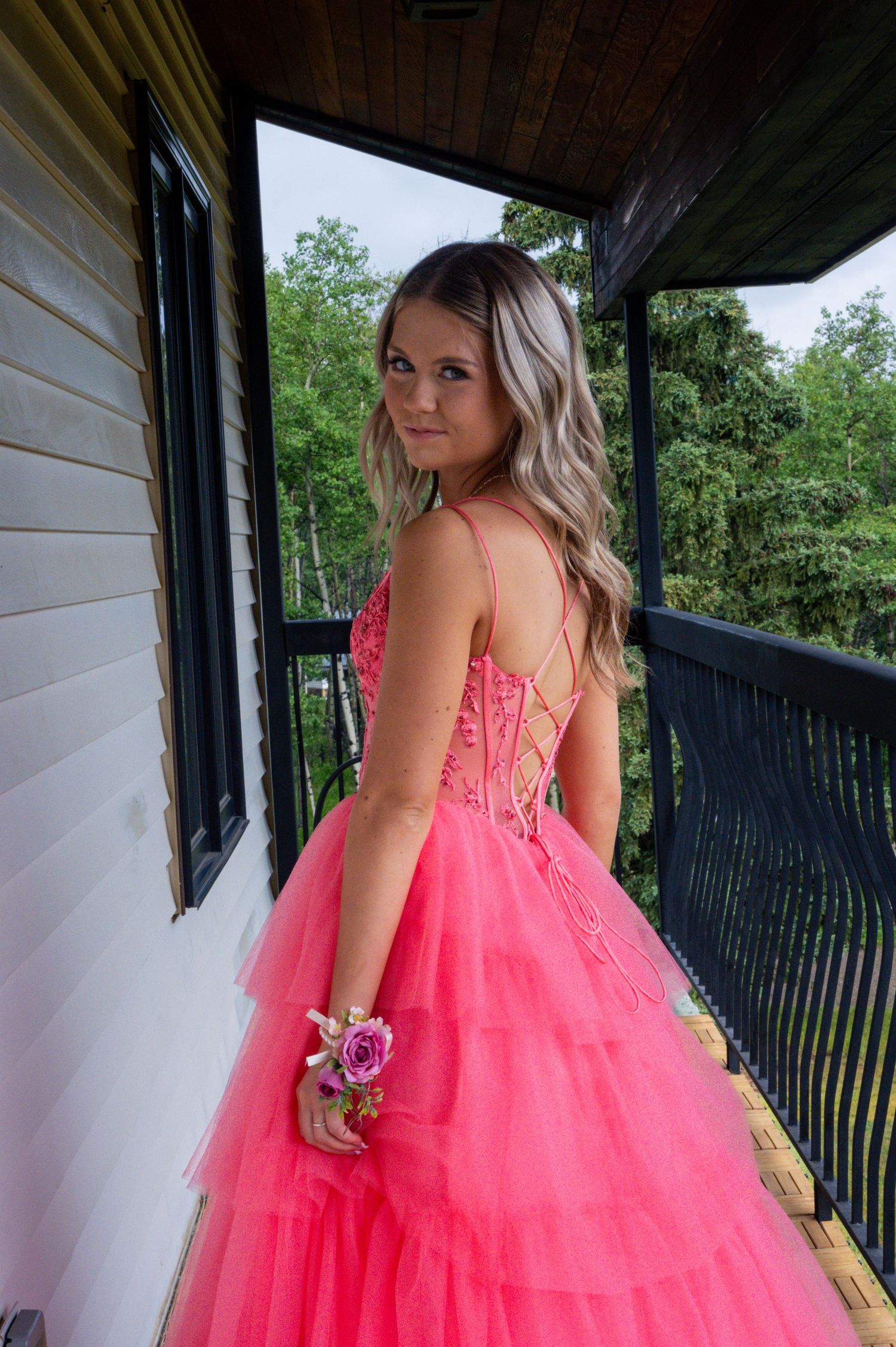 Person in a stunning coral pink layered tulle prom gown with lace corset detail poses on a rustic cabin porch.
