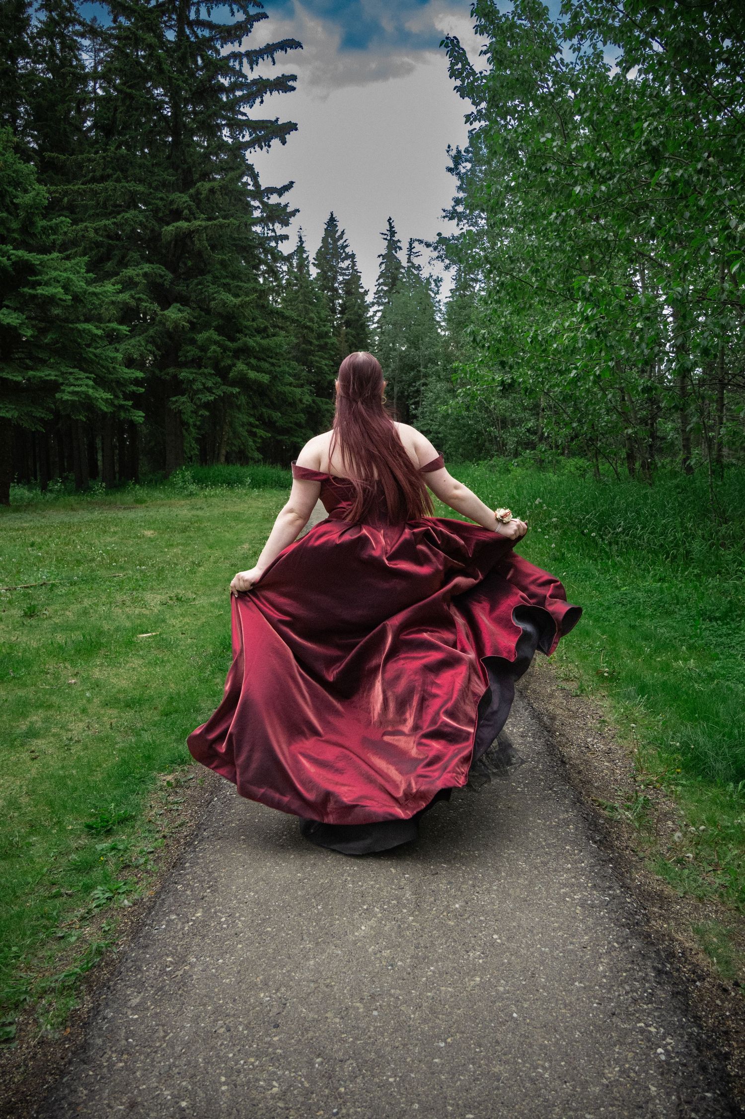 A burgundy dress flows dramatically as someone walks down a forest path surrounded by evergreen trees.