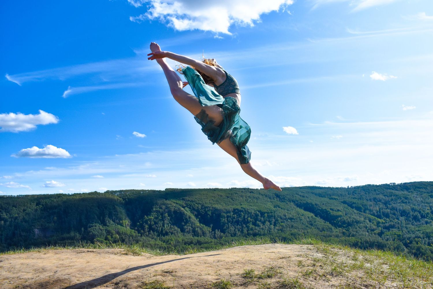 A dancer leaps gracefully through the air against a backdrop of mountains and blue sky.