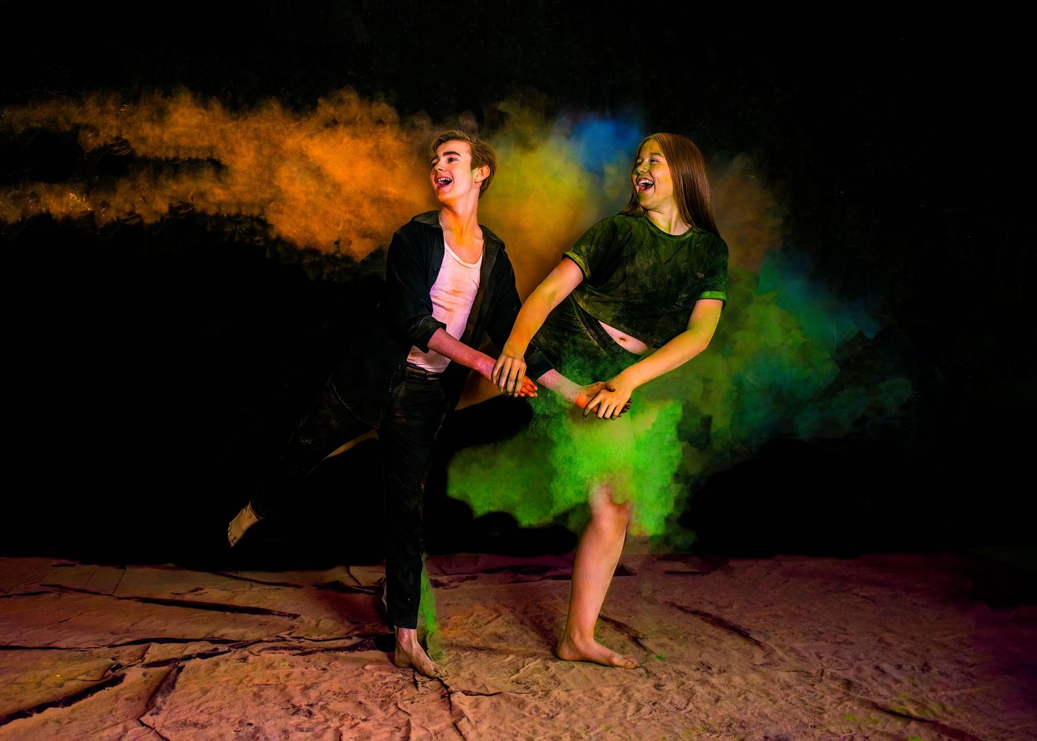 Dancers performing amid swirling orange and blue powder effects on a dark stage.