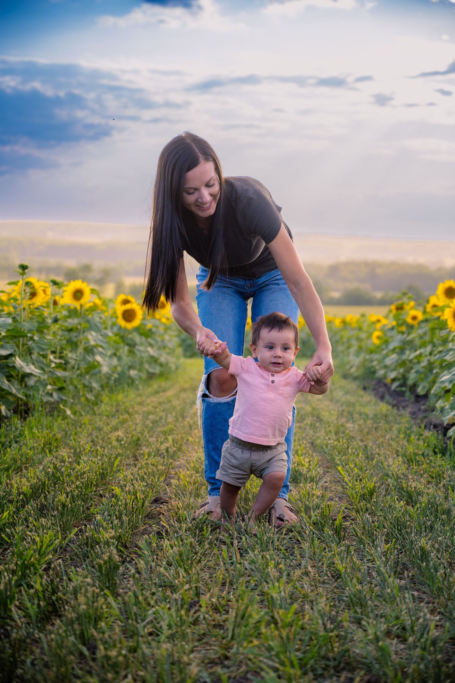 Parent helping toddler take first steps in sunflower field during sunset.