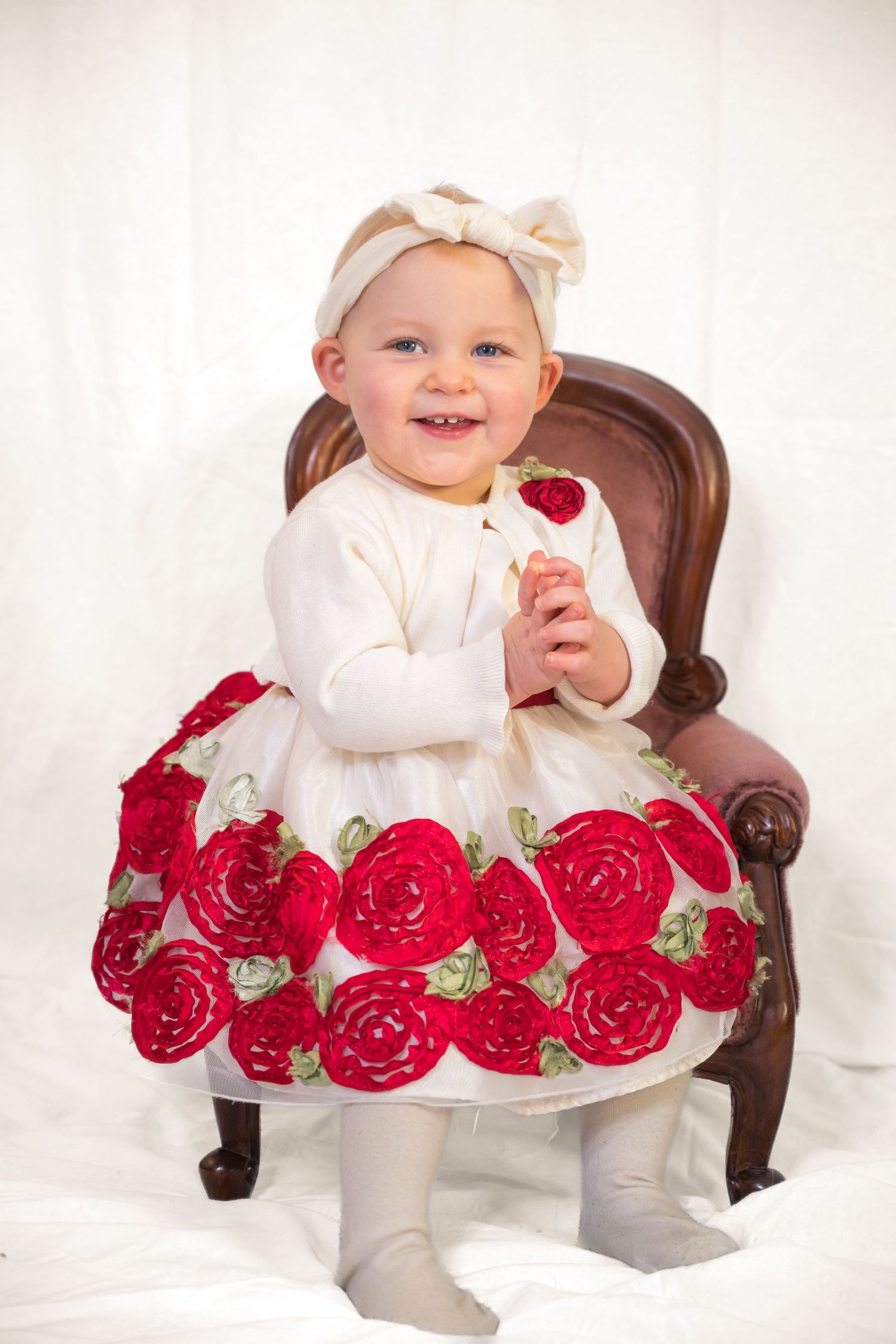 Small child in white dress with red rose pattern sits in vintage brown chair.