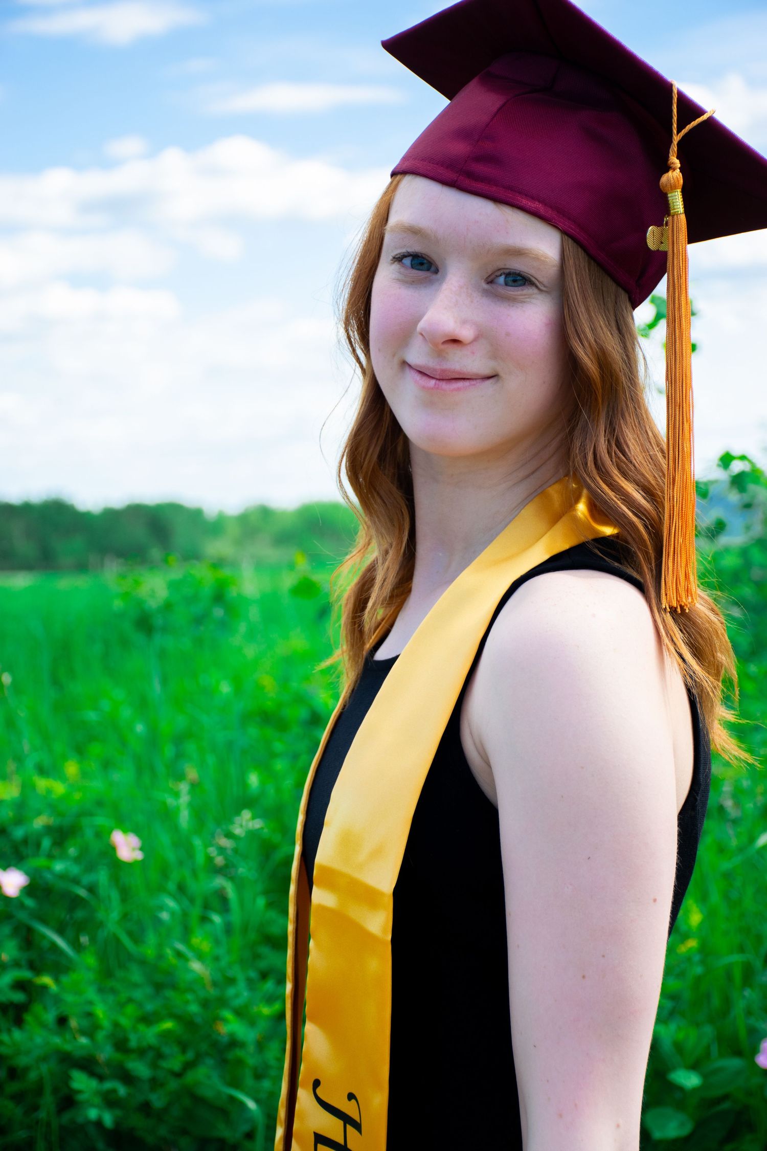 Graduate in burgundy cap and gold sash poses in sunny field.