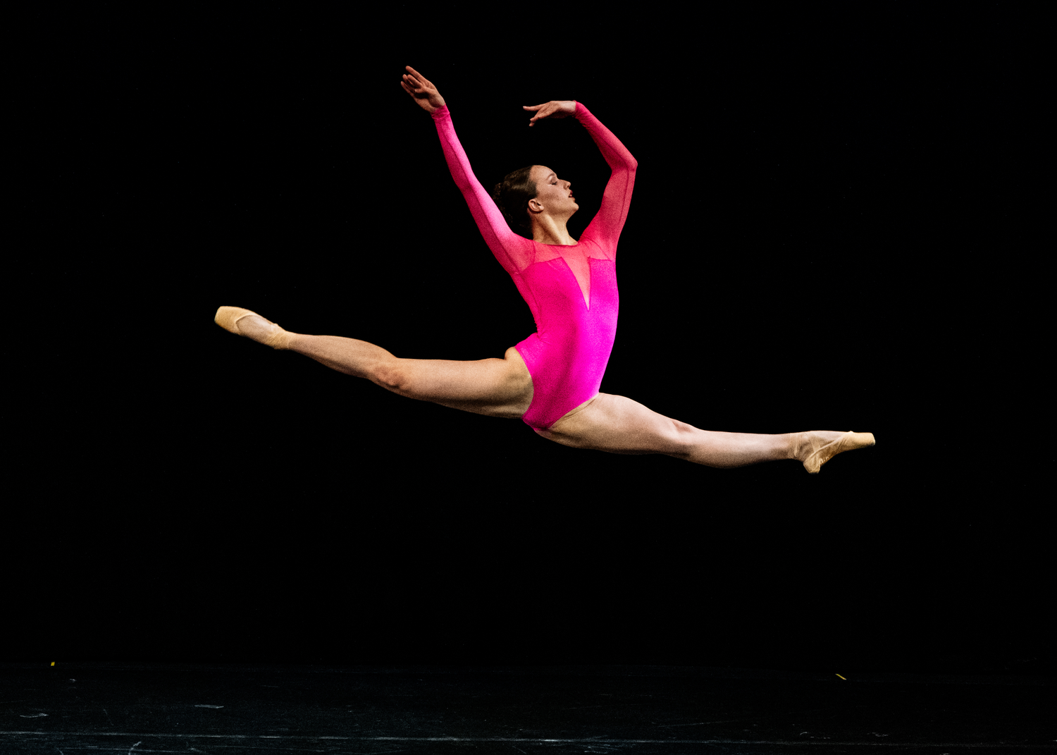 Dancer in pink leotard performs dramatic split leap against black background.