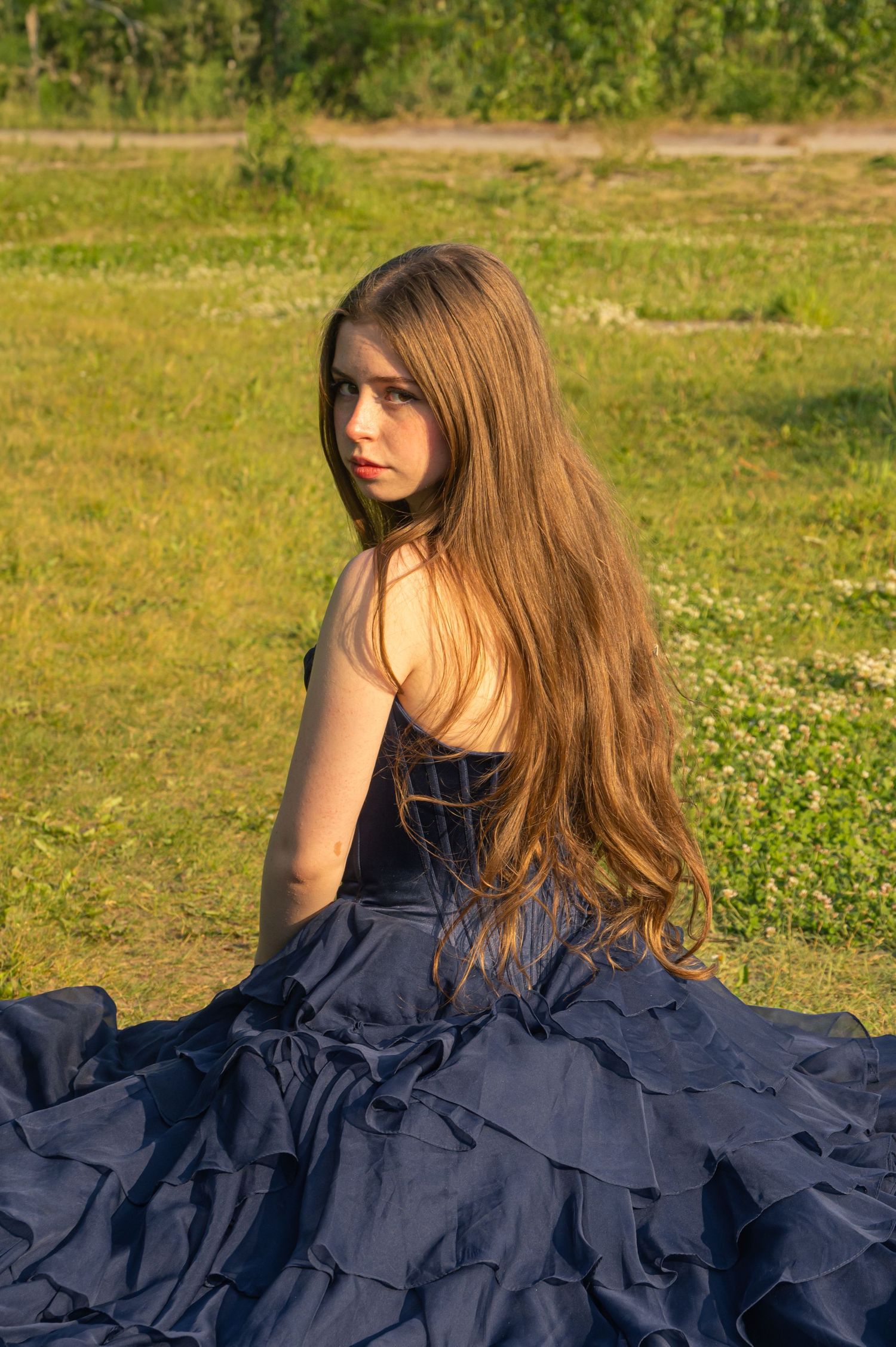 A person in an elegant navy blue ruffled dress sits in a grassy field on a sunny day.