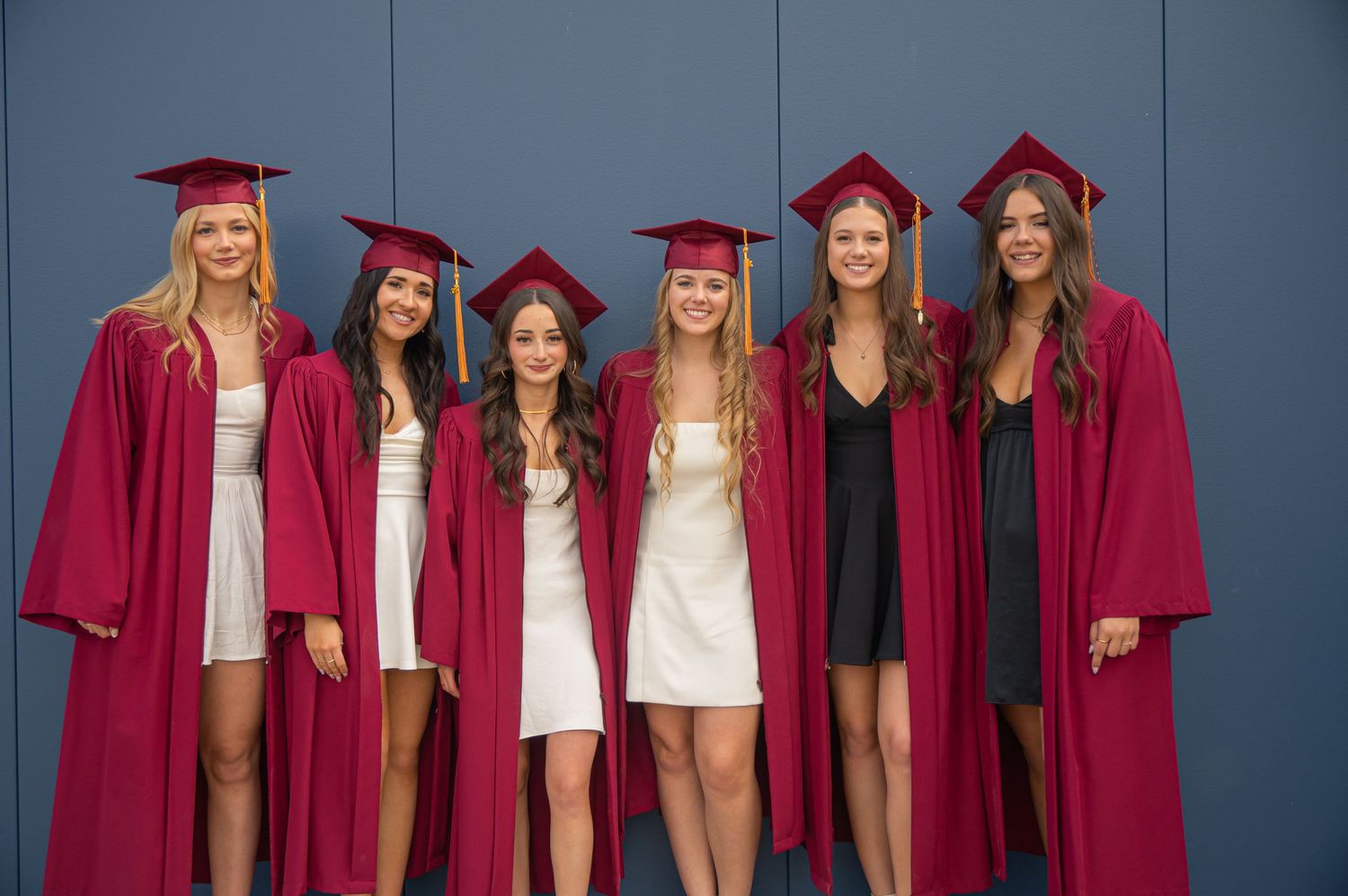 A group of graduates wearing crimson red gowns and caps stand together against a blue background.