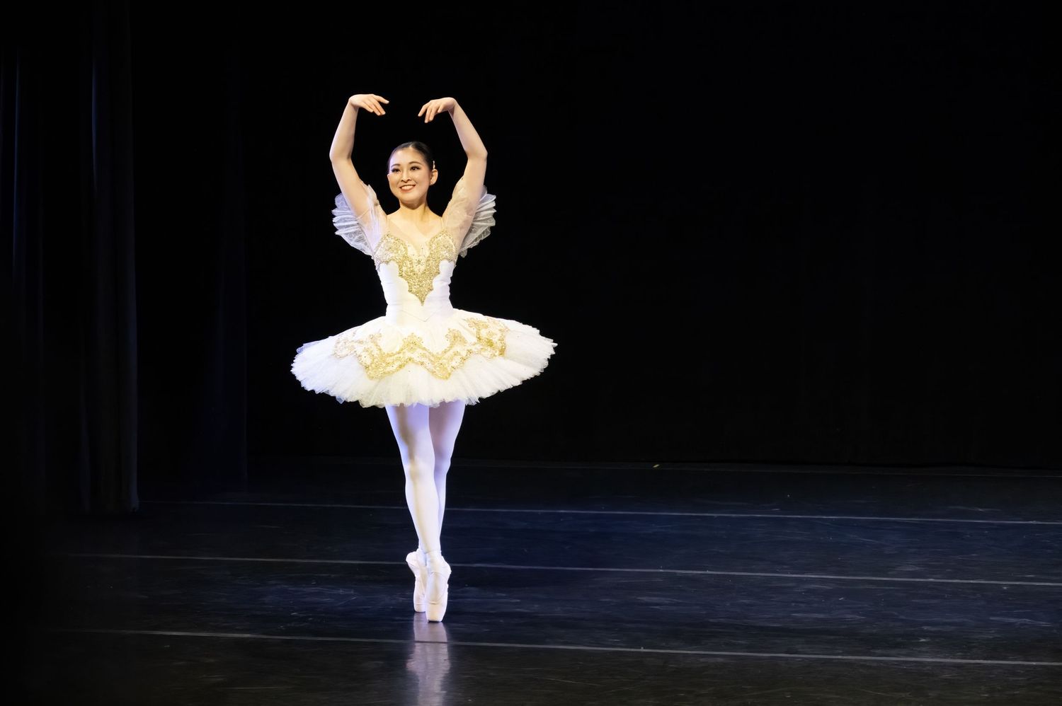 A ballet dancer in a white tutu performs on point against a dark stage background.