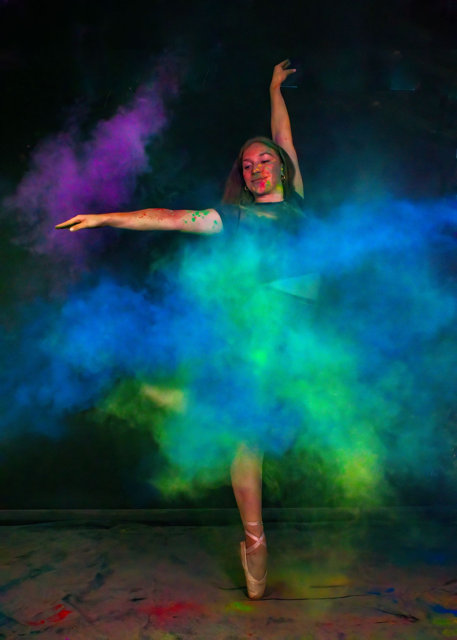 Ballet dancer in pointe shoes performing amid swirling blue and green powder clouds.