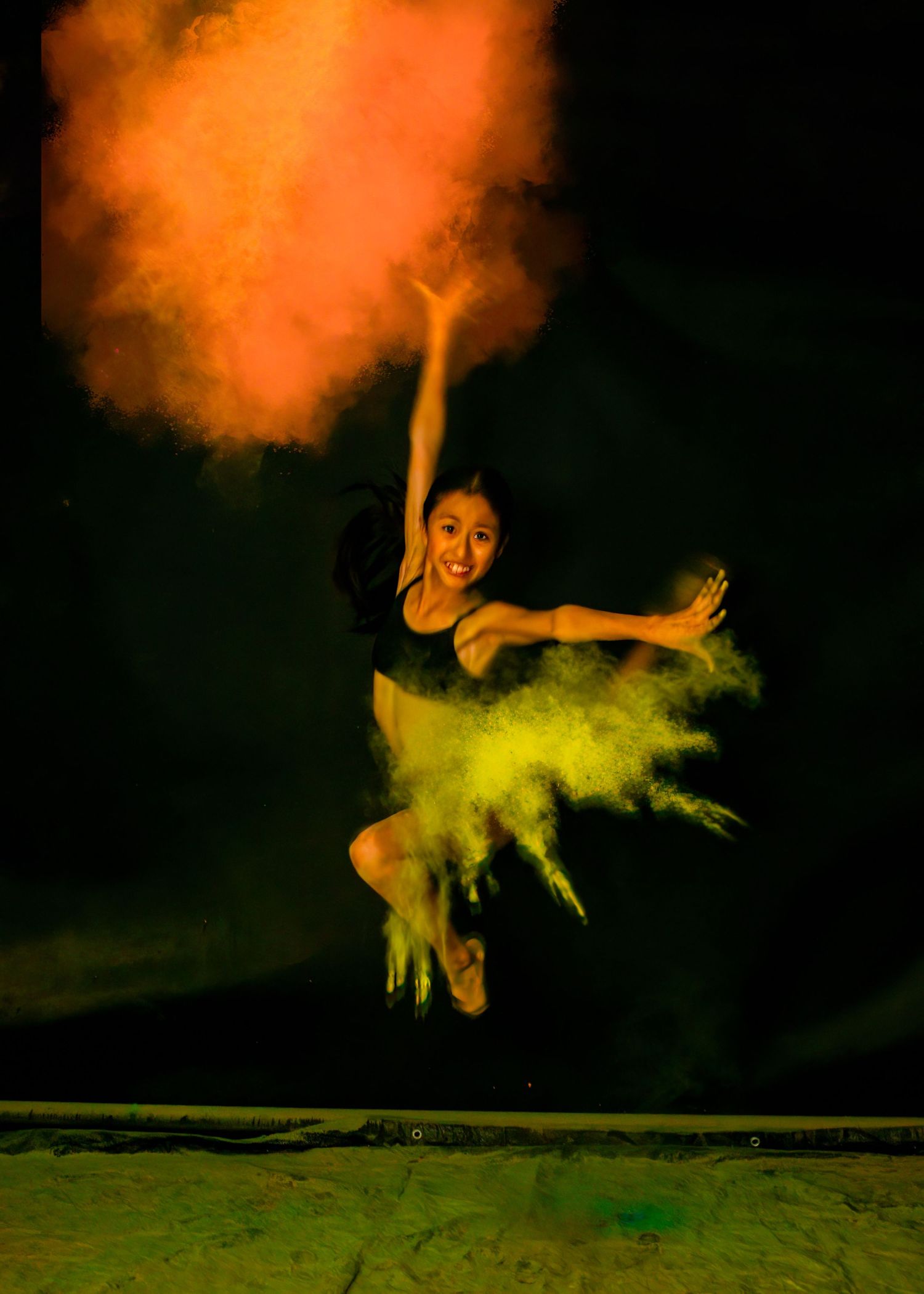 Dancer leaping through vibrant orange and yellow powder clouds against dark background.