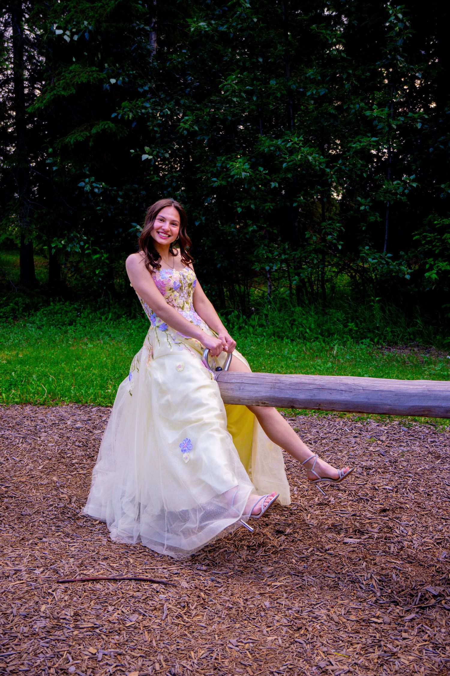 A bride in a white wedding gown sits on a wooden bench in a lush garden setting on a bed of mulch.