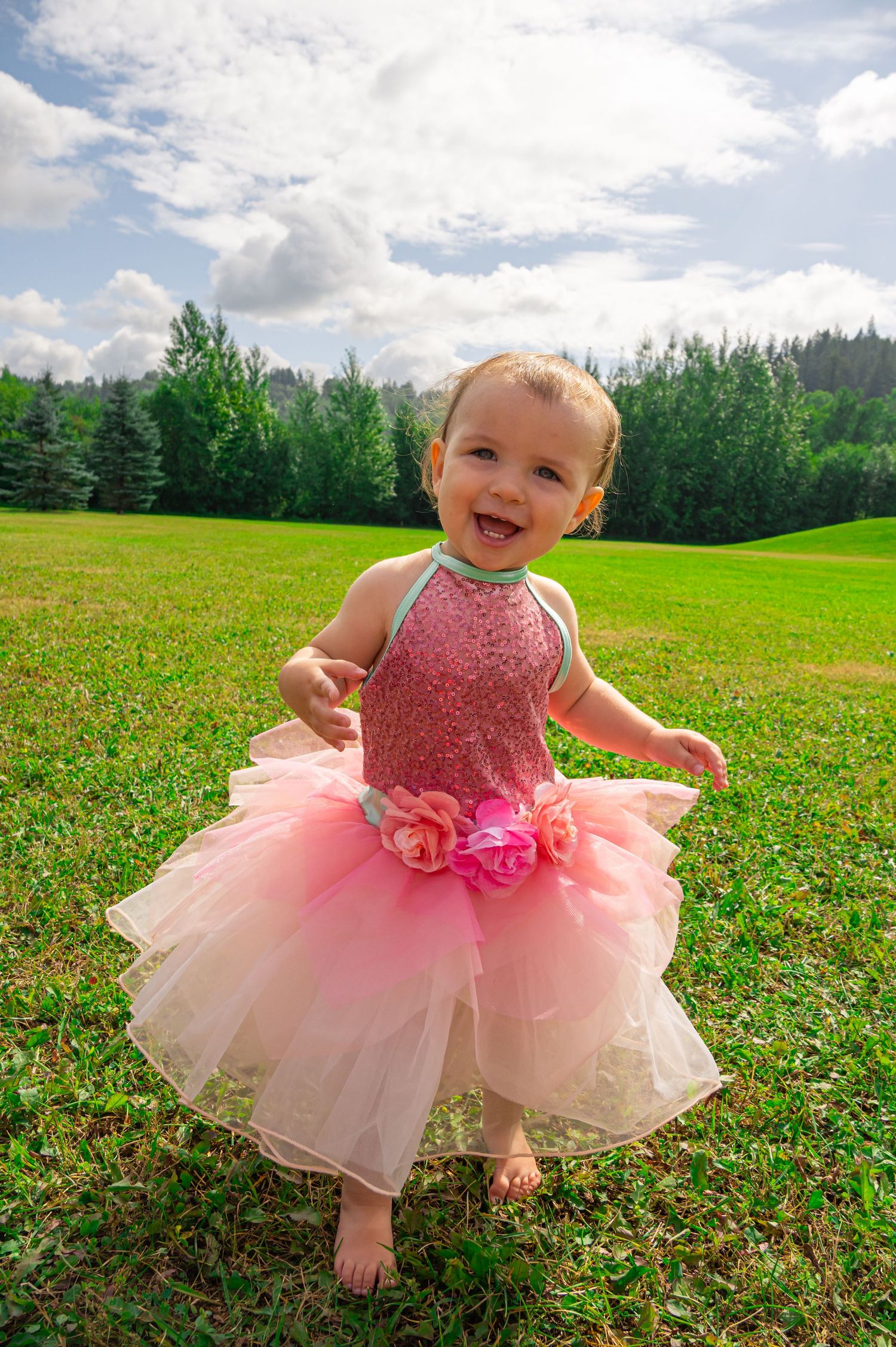 A pink sparkly tutu dress with floral details twirls on a sunny day against a backdrop of evergreen trees.