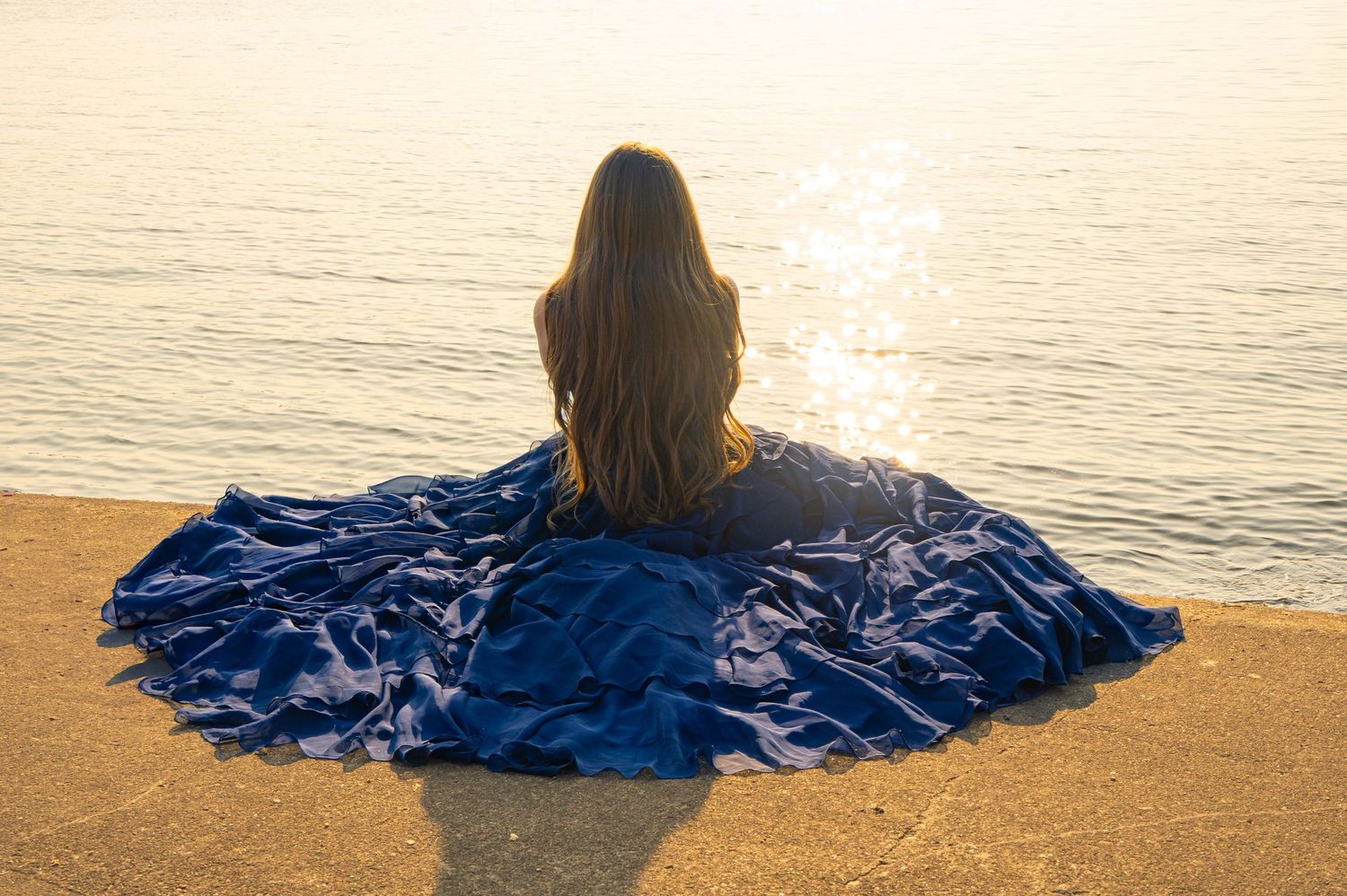 Figure in flowing blue dress sits on beach watching golden sunset over ocean waters.