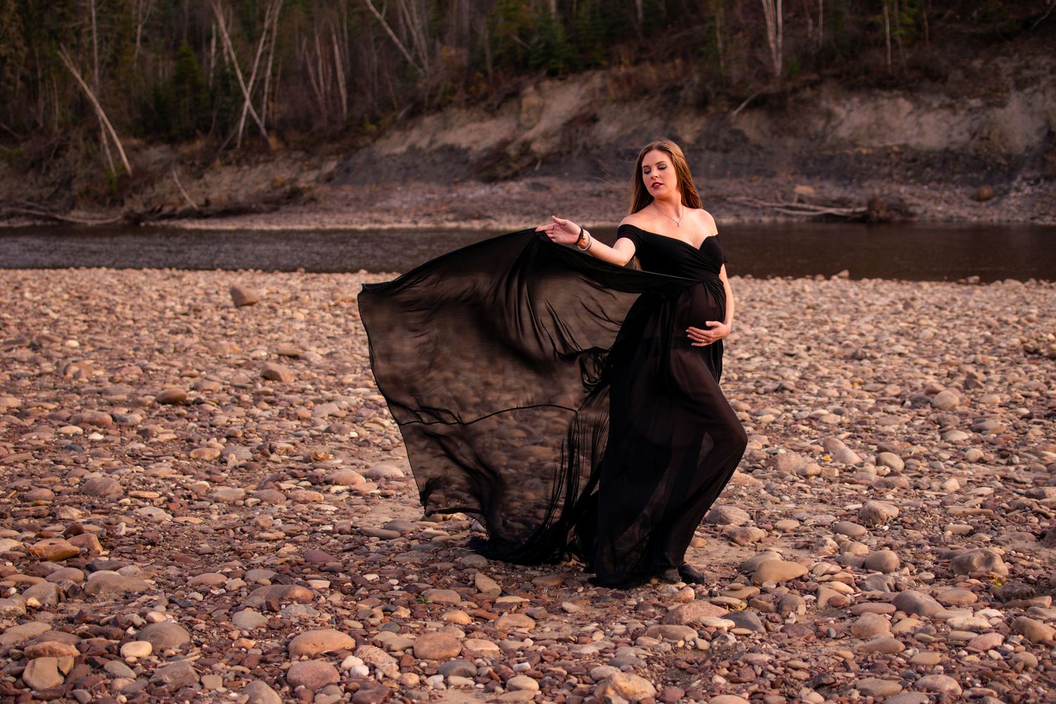 A flowing black dress billows dramatically as someone poses on a rocky riverbed at sunset.
