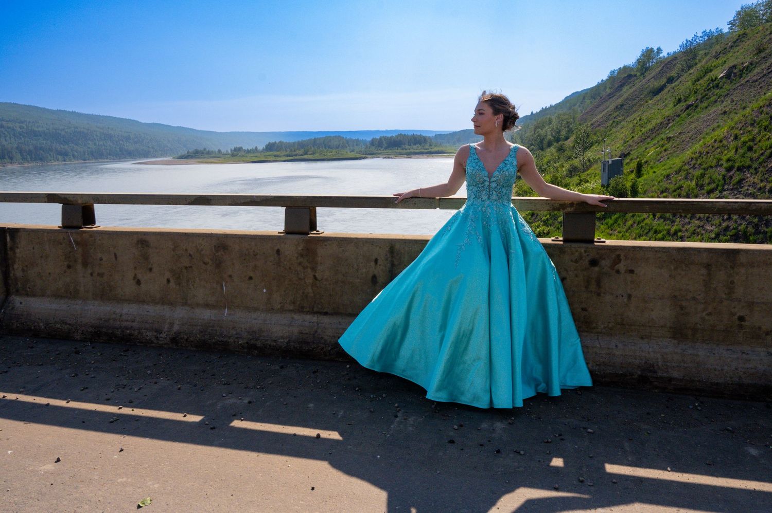 Someone in a flowing turquoise formal dress poses by a stone wall overlooking a scenic mountain lake on a sunny day.