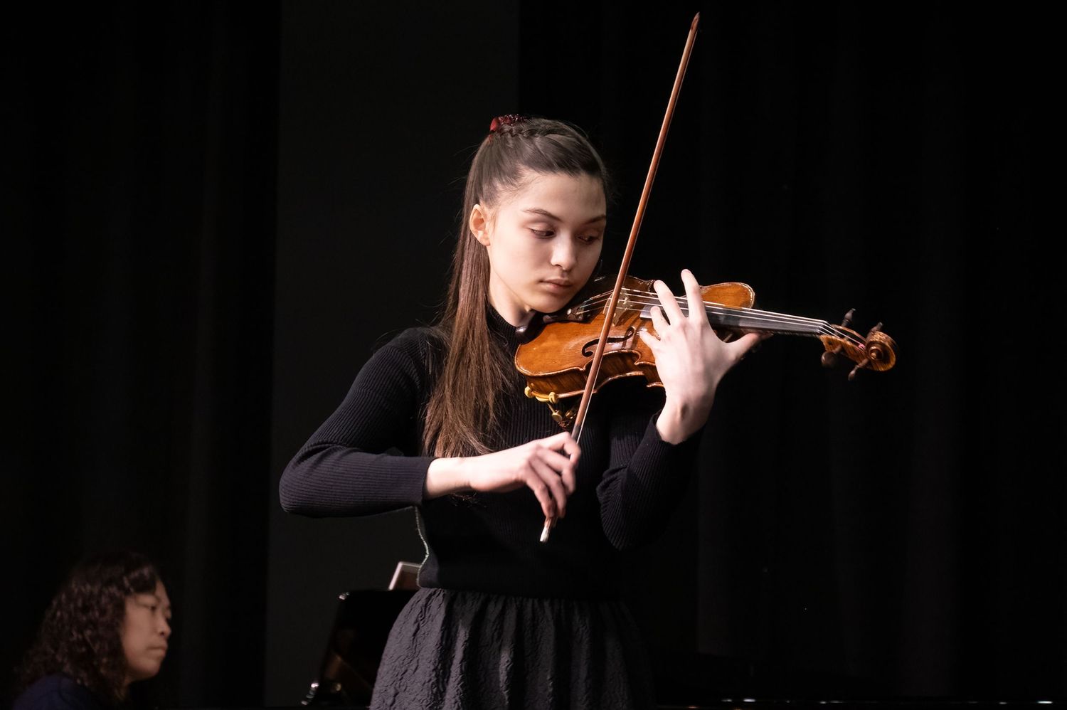 Musician in black dress performing on violin against dark background during classical concert performance.