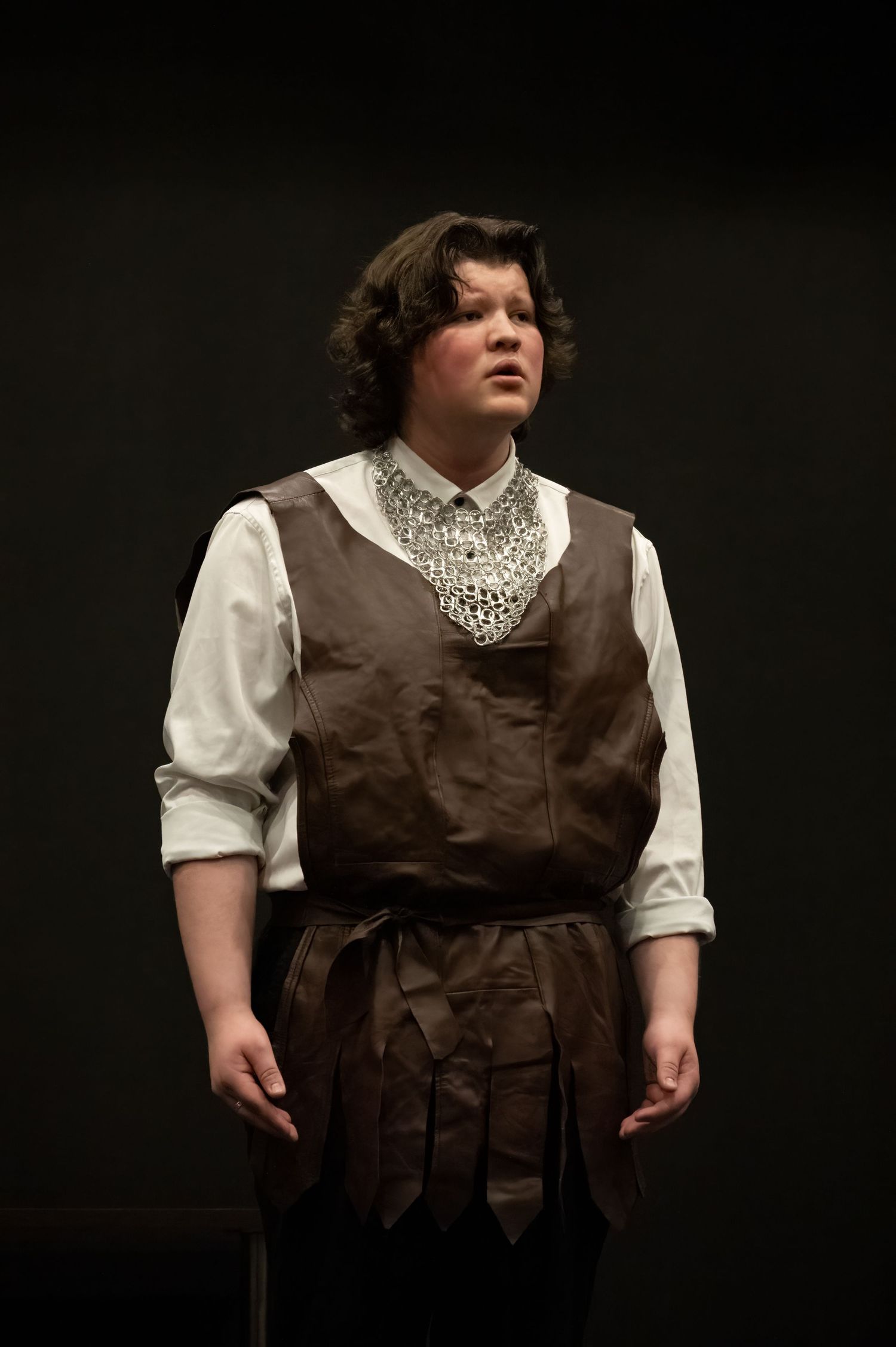 A performer in a period costume consisting of a brown vest, white shirt with lace collar stands against a dark background.