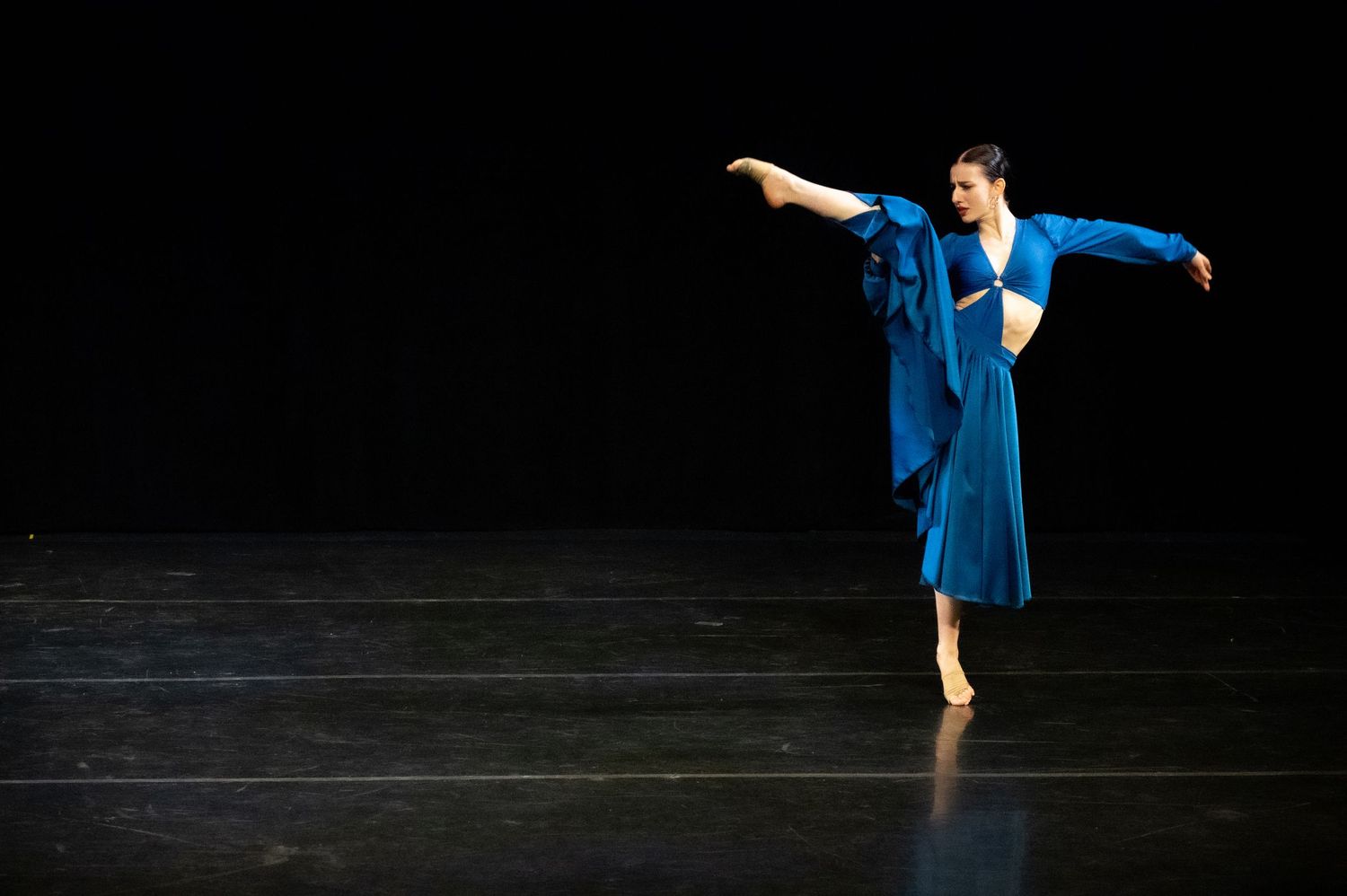 Modern dancer in flowing blue dress performs graceful pose on dark stage during contemporary dance performance.