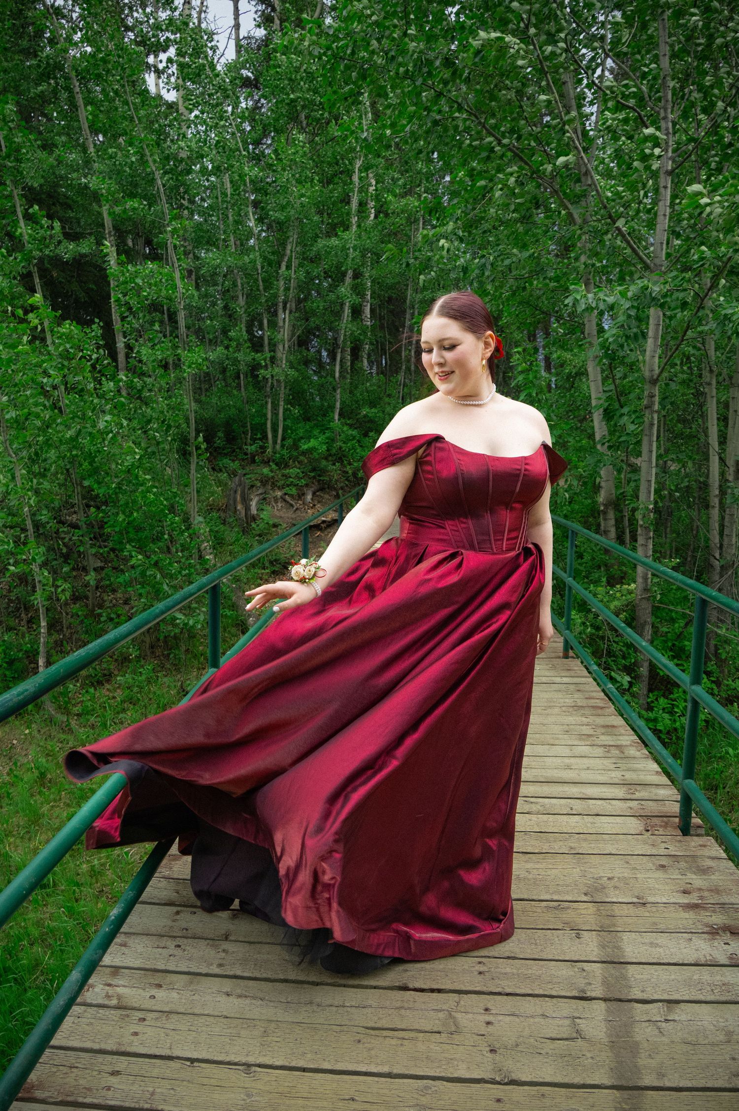 A person in a flowing burgundy gown twirls playfully on a wooden footbridge surrounded by lush green forest.