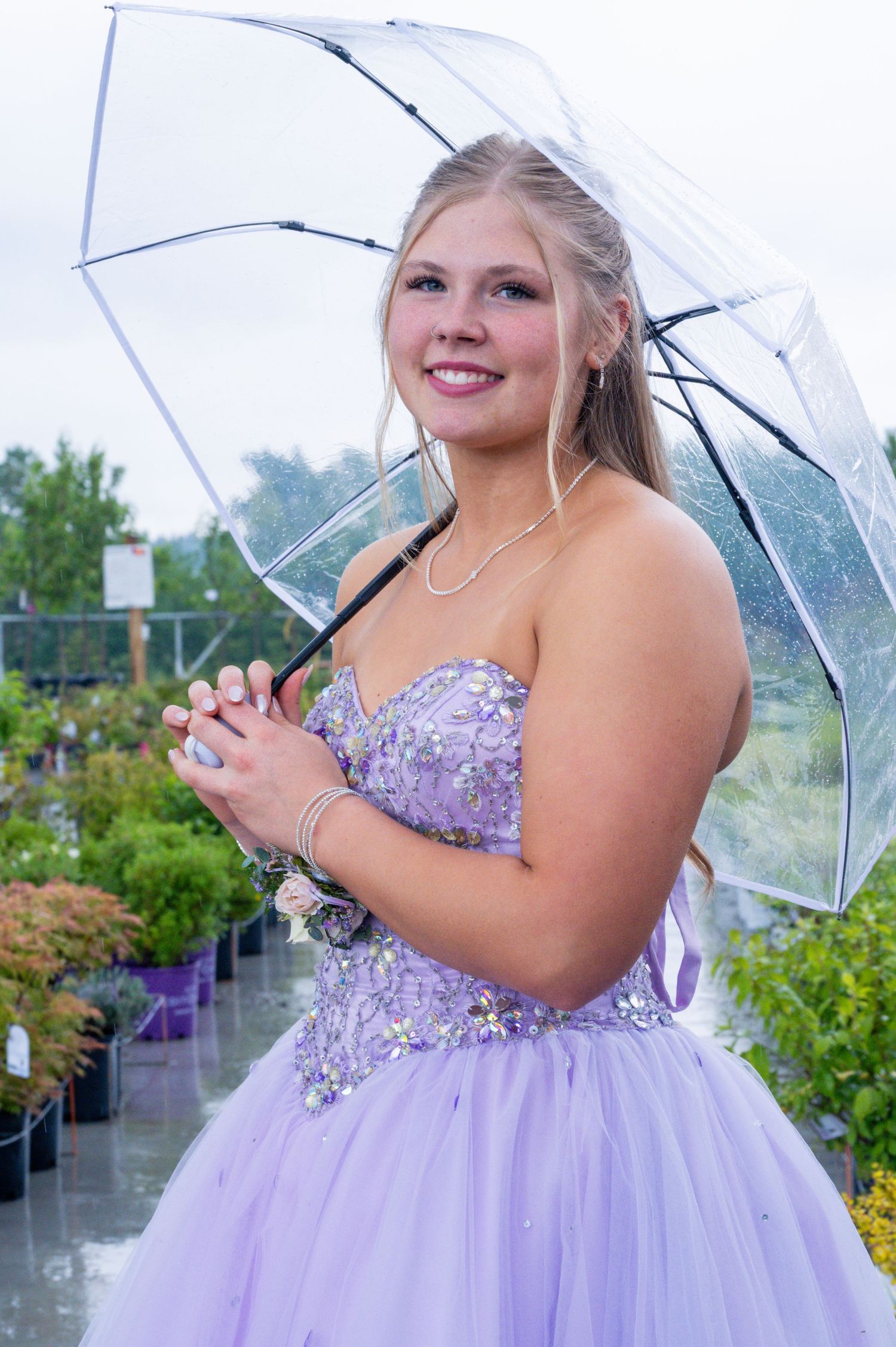 A person in a lavender prom dress holds a clear umbrella while standing in a garden on a rainy day.