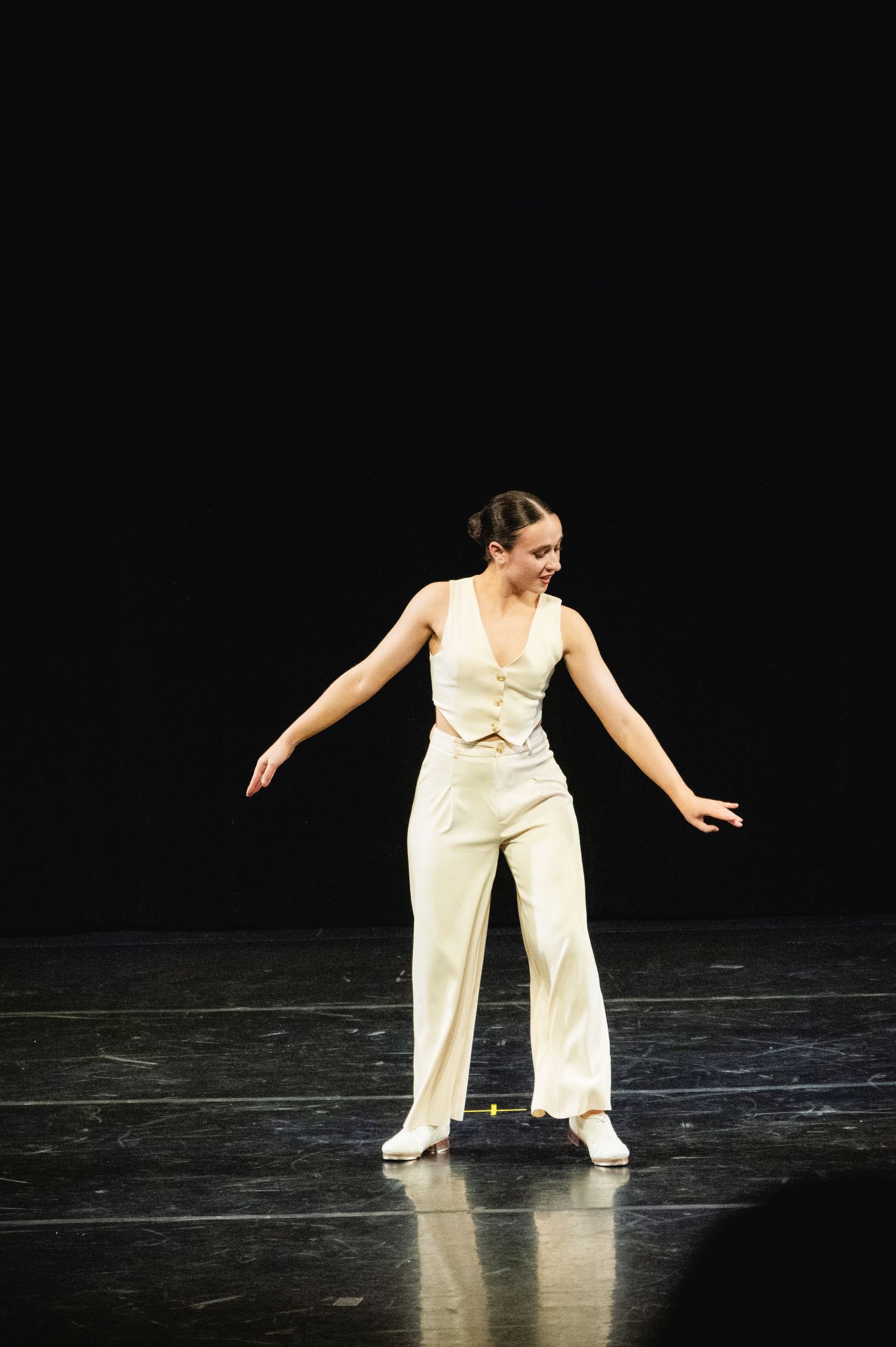 A dancer in a cream-colored jumpsuit performs on stage against a dark background, arms outstretched in graceful pose.
