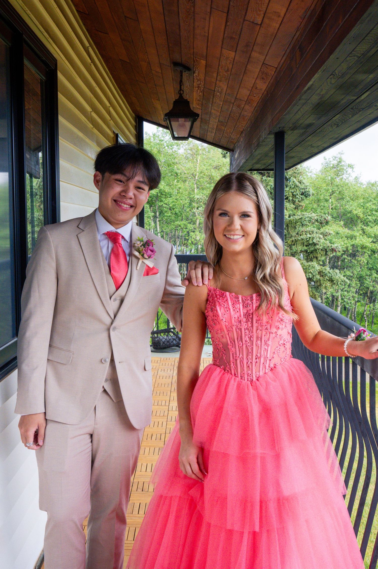 Two formal event attendees in coordinating pink and beige outfits pose together on a covered porch.
