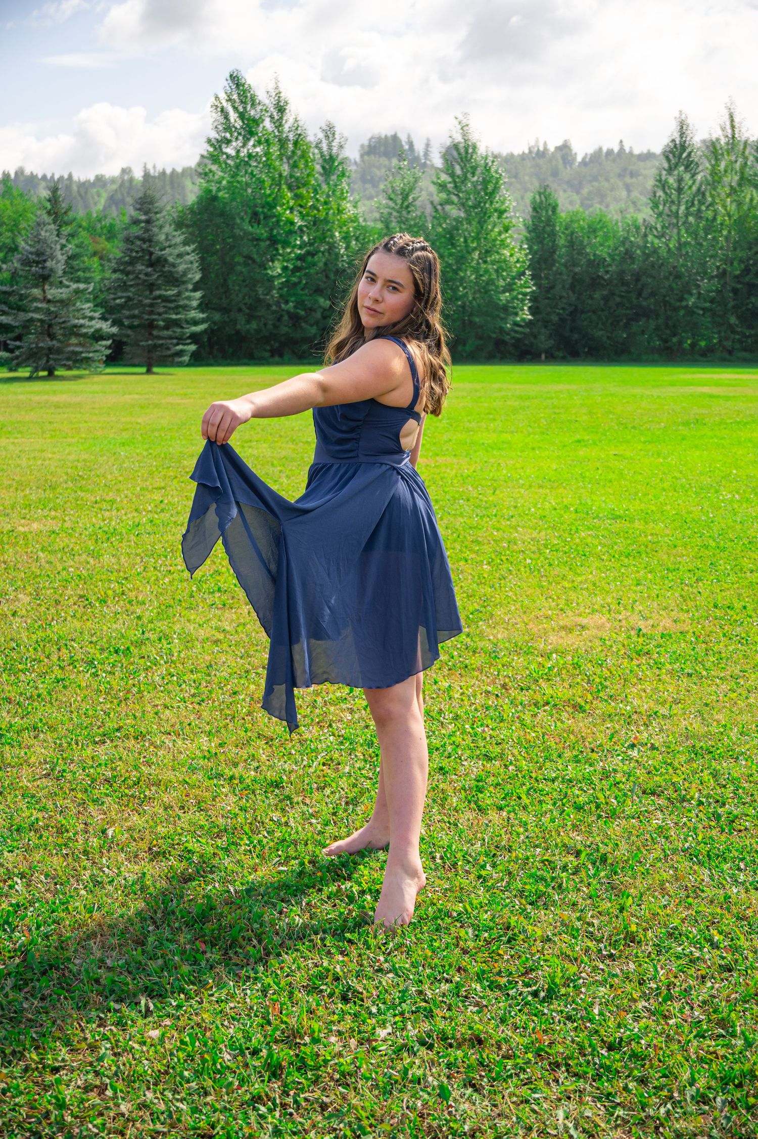 A navy blue dress flows gracefully as someone twirls on a lush green lawn with pine trees in the background.