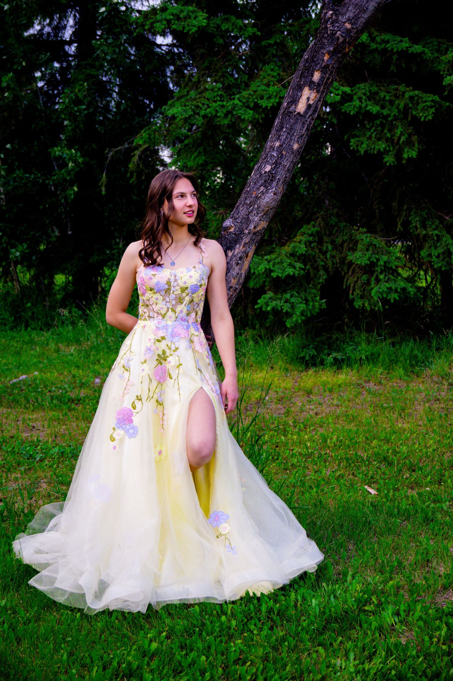 Woman wearing a floral patterned white and yellow ballgown poses gracefully in a lush green outdoor setting.