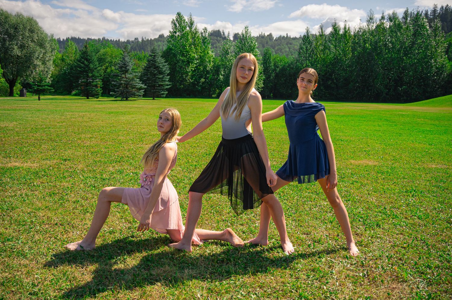 Three dancers strike graceful poses on a sunny green lawn surrounded by evergreen trees in a park setting.