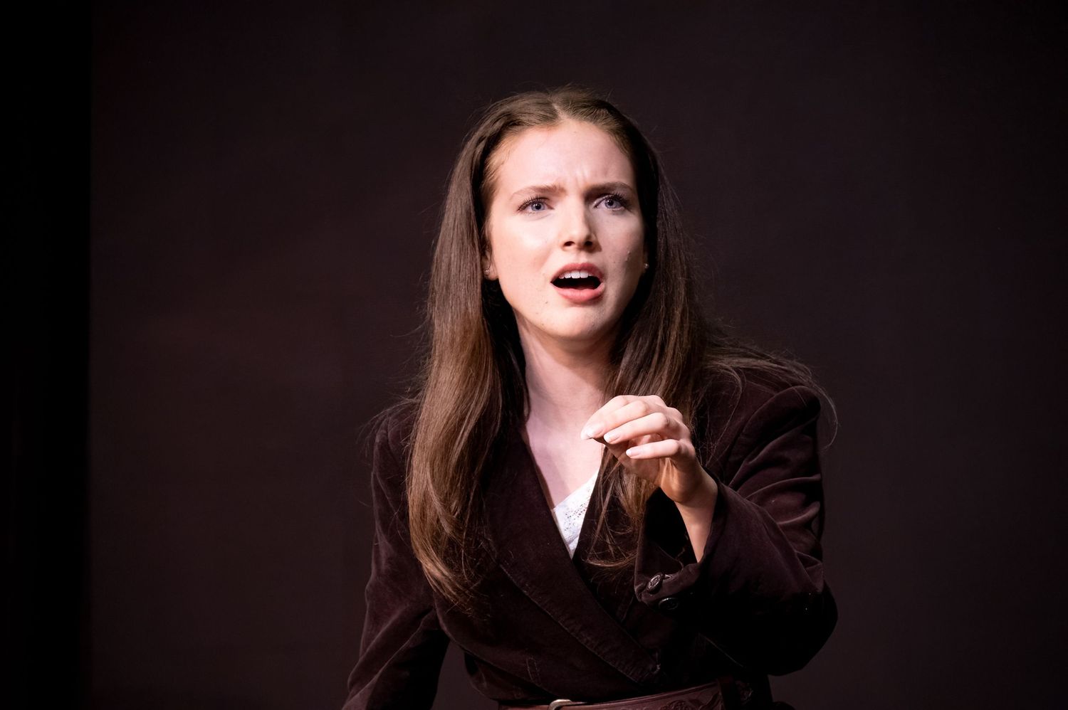 Person in brown blazer gestures expressively against dark background while performing or speaking.