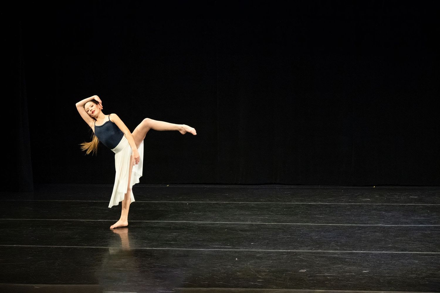A dancer performs a graceful arabesque pose in a white costume against a dark stage background.