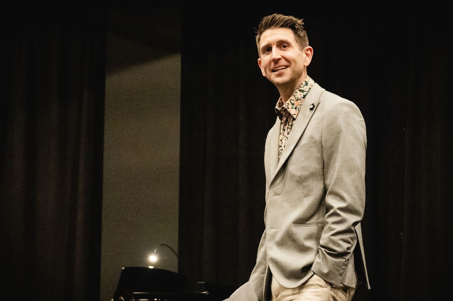 A person wearing a beige suit jacket stands on stage in dramatic lighting with dark curtains in the background.