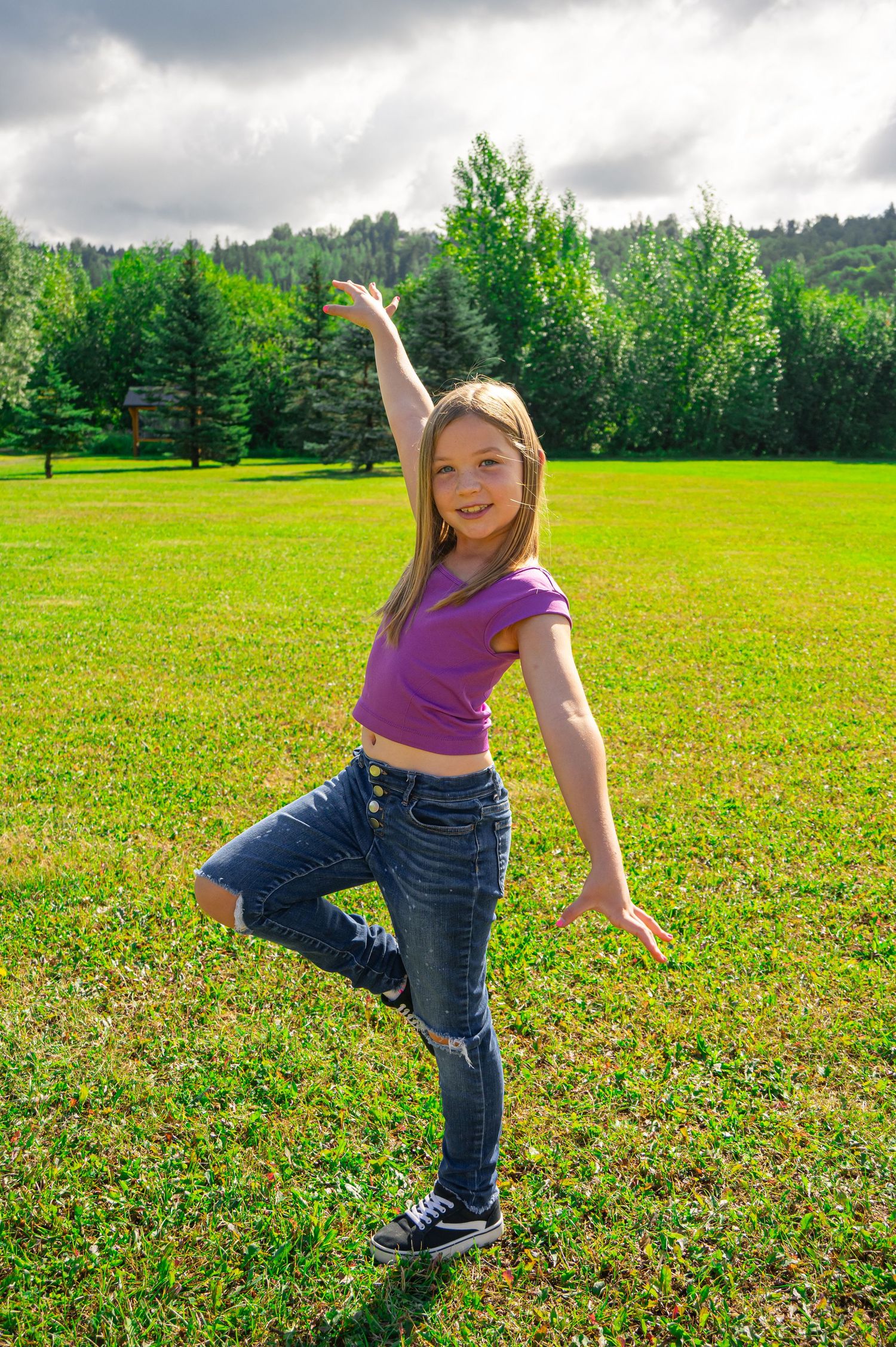 Someone strikes a playful pose in a purple crop top and jeans on a sunny grassy field with trees in the background.