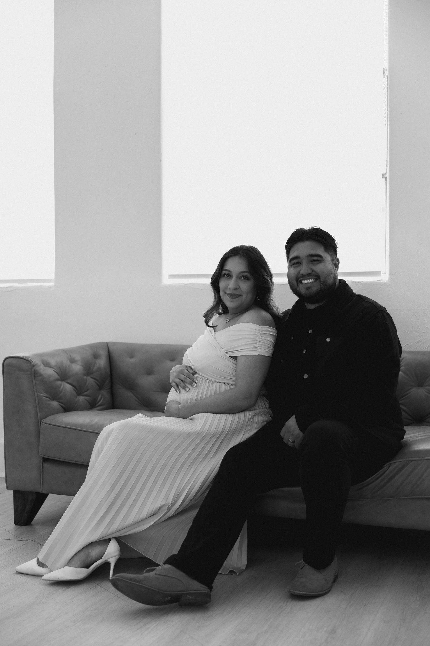 A couple sits together on a gray couch in a minimalist room with white walls, captured in black and white.