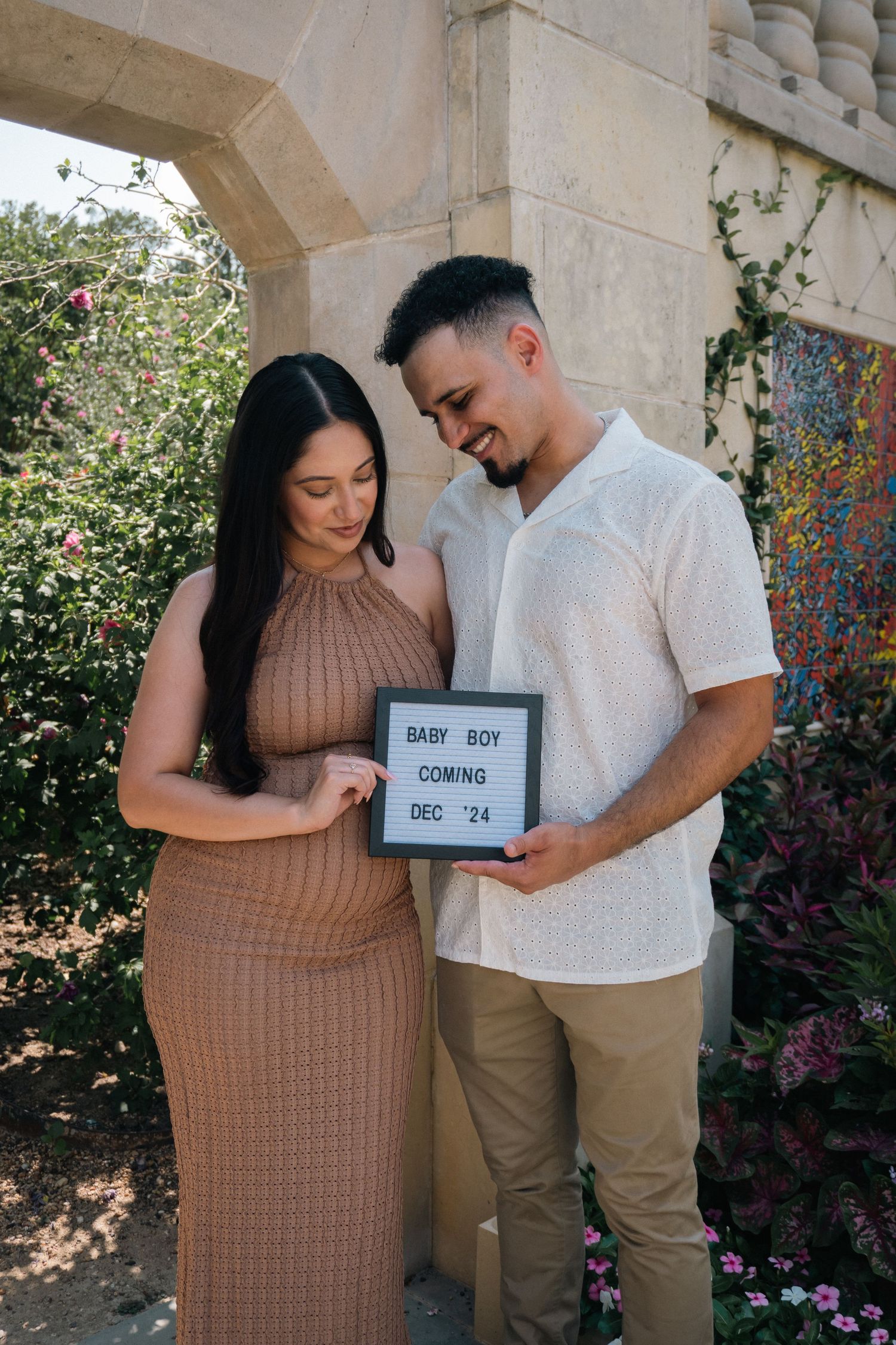 Couple shares pregnancy announcement with letter board signs while standing together in outdoor garden setting.