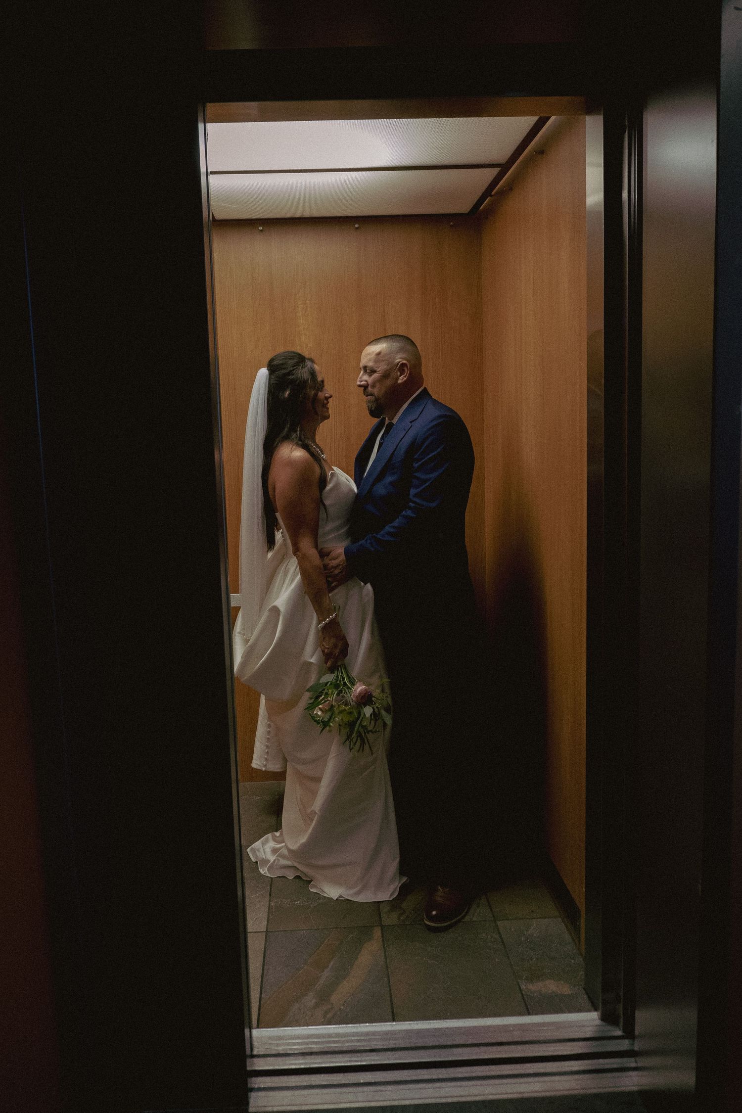 A couple shares an intimate moment in an elevator, with one person wearing a white dress and holding a bouquet.