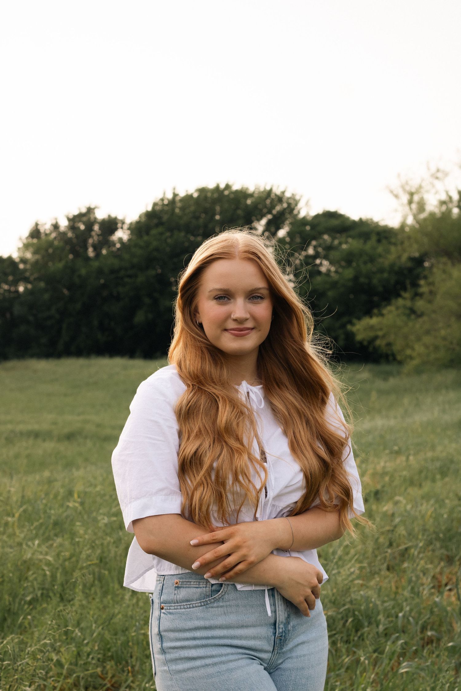 A person with long red hair poses in a white t-shirt and jeans against a natural outdoor backdrop at sunset.