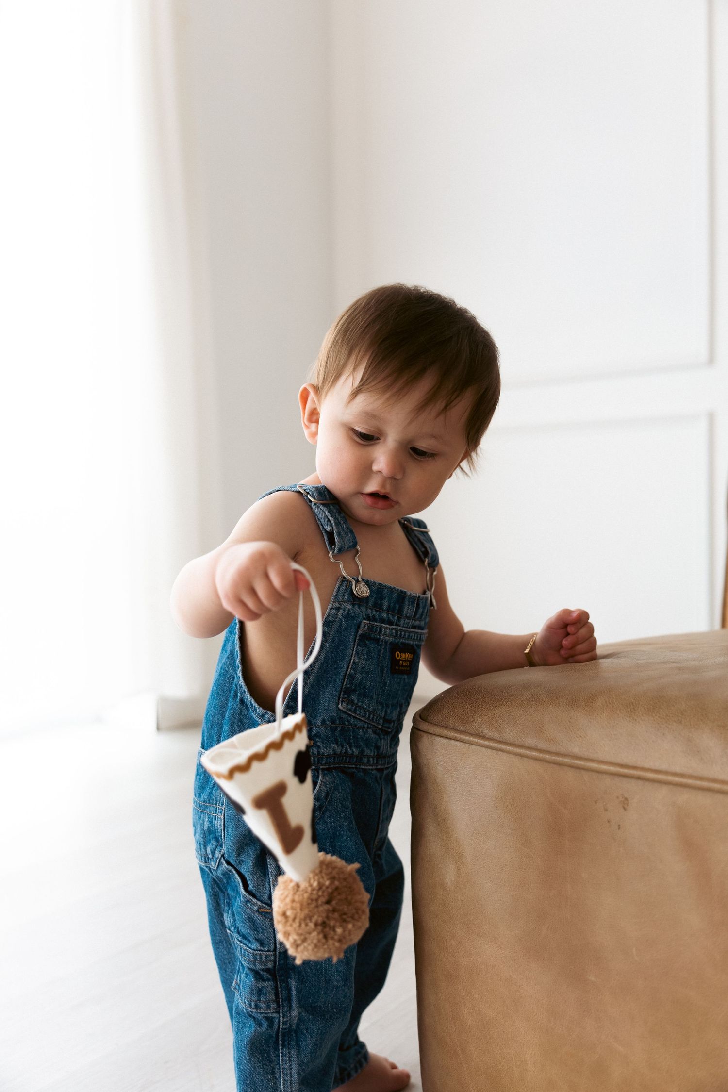 Small child in denim overalls plays near a beige couch in a bright, minimalist room.