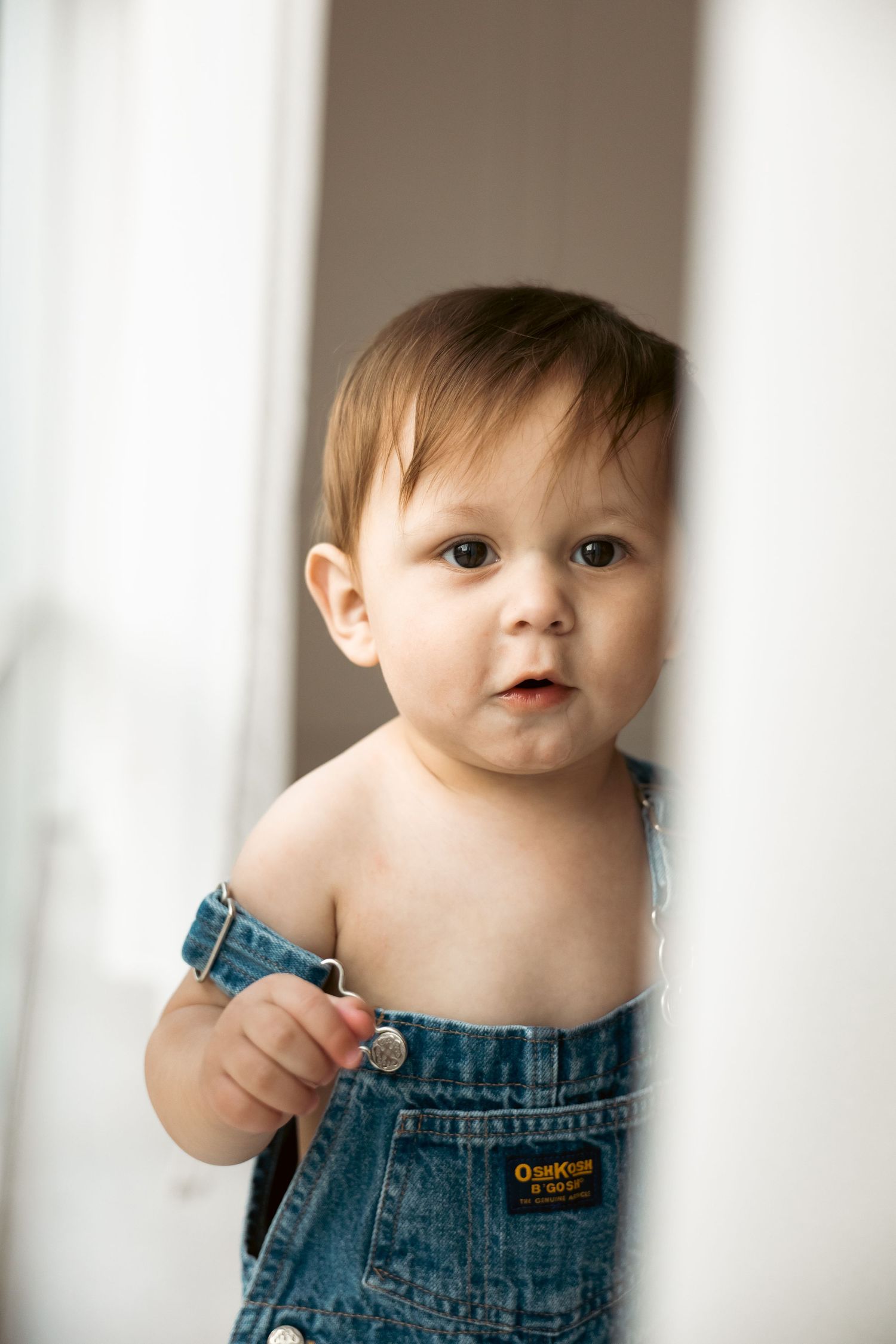 Sweet baby in denim overalls poses against white background in soft natural light.