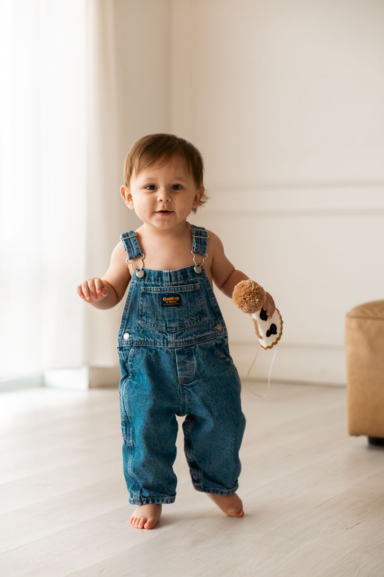 A toddler in denim overalls holds cookies while walking on a light wooden floor in a bright minimalist room.