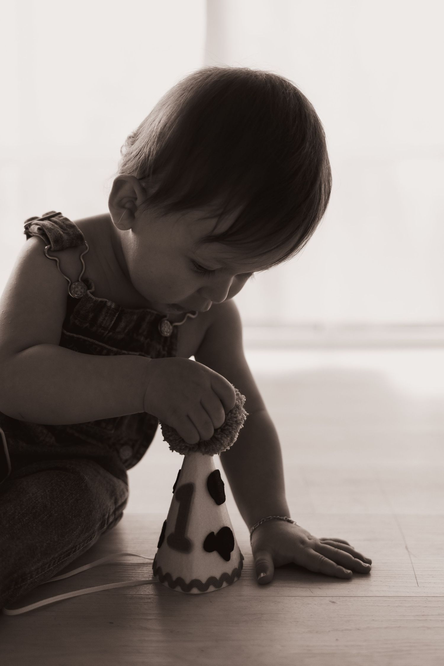 A young child in overalls plays with a small ghost-shaped decoration on a wooden floor in a sepia-toned photograph.