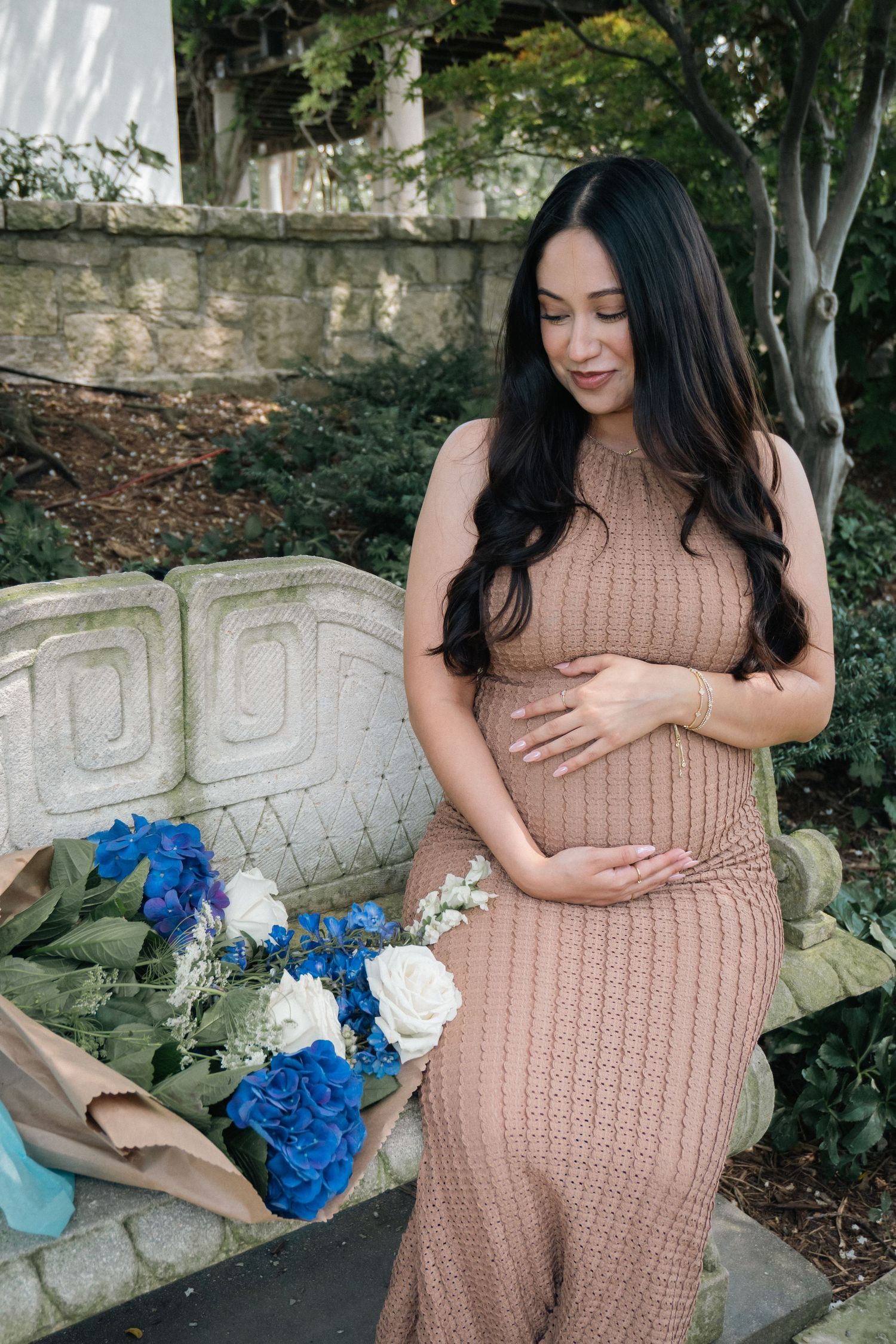 A maternity photo series showing a woman in a beige knit dress holding her baby bump in a garden setting.
