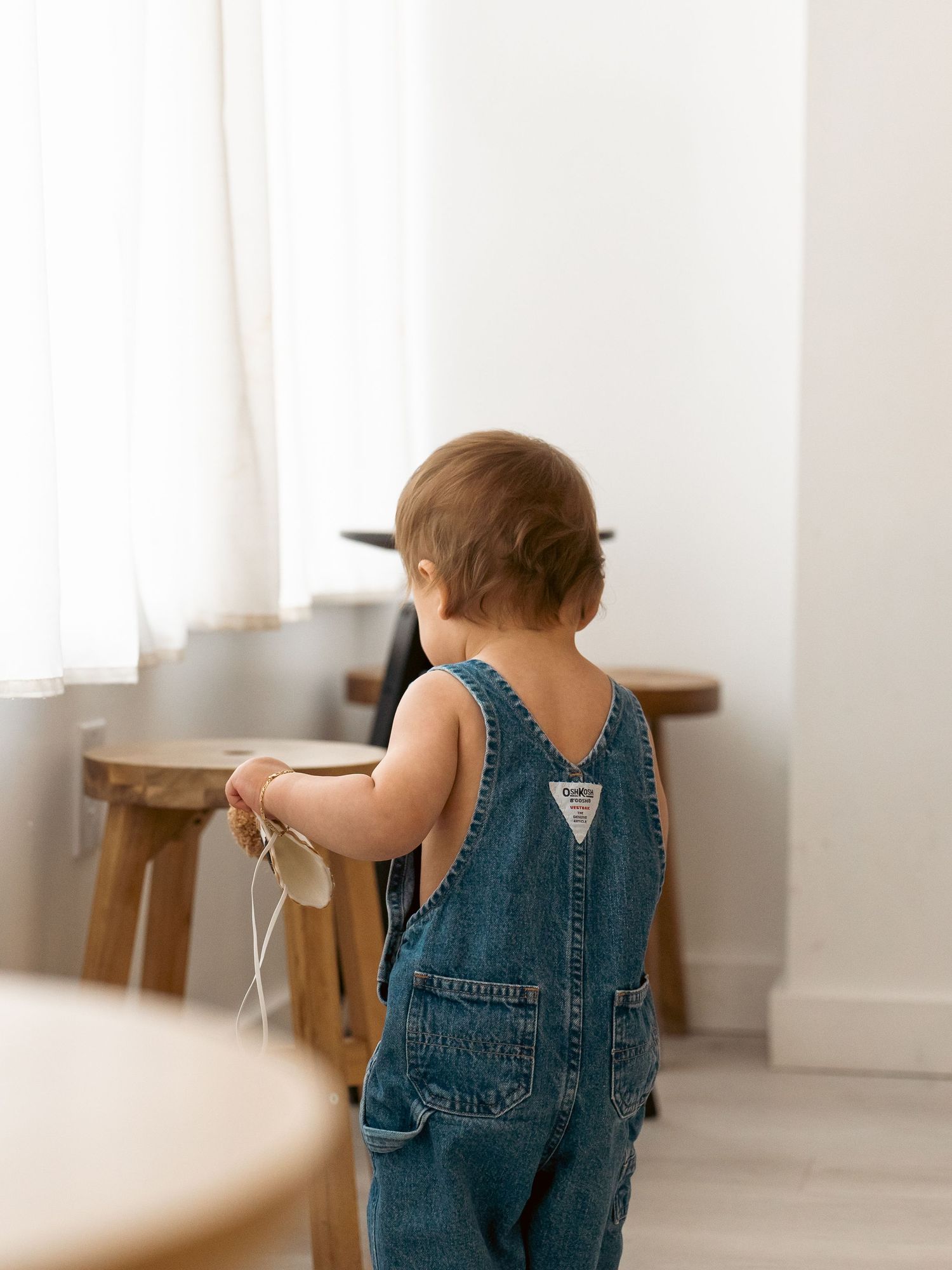 Toddler wearing blue denim overalls walks through bright minimalist room with wooden furniture.