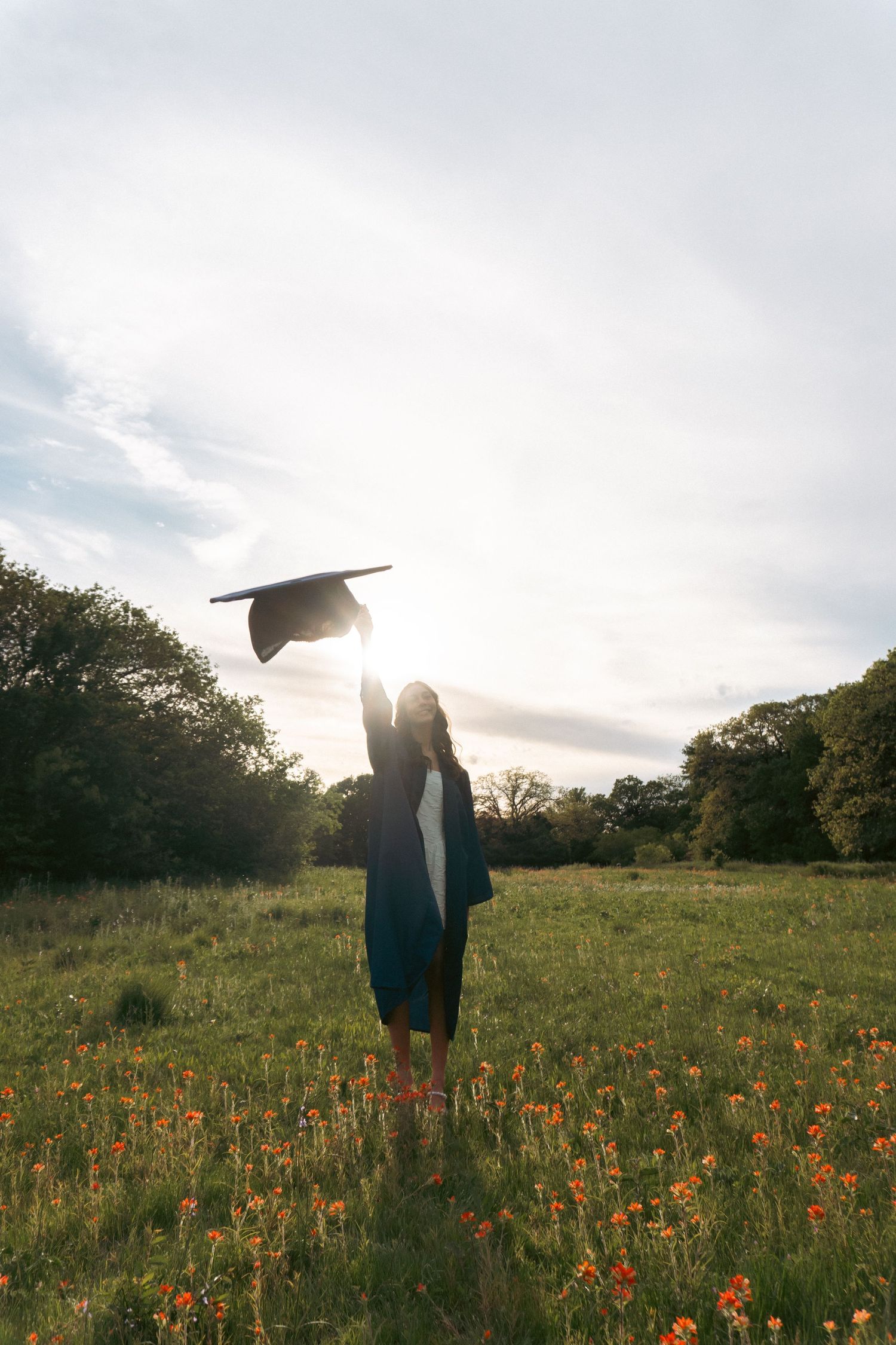 A graduation cap is tossed in the air against sunlit sky in a wildflower meadow during golden hour.
