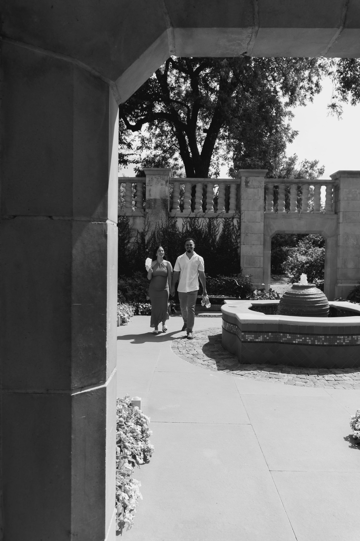 A couple strolls through an elegant garden courtyard with stone balustrades and a fountain in black and white.