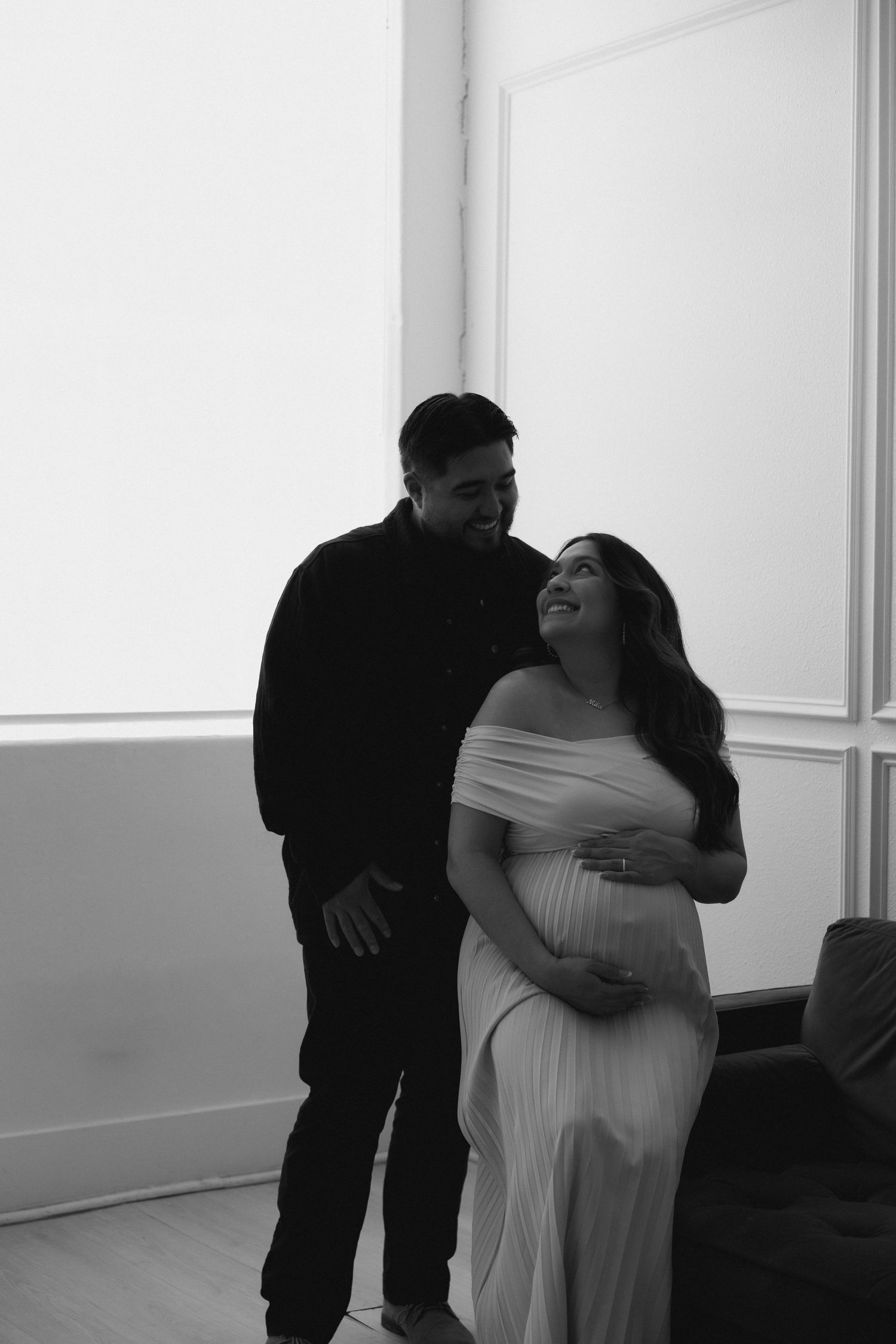 Black and white maternity photo showing a couple sharing a joyful moment together in a bright indoor setting.