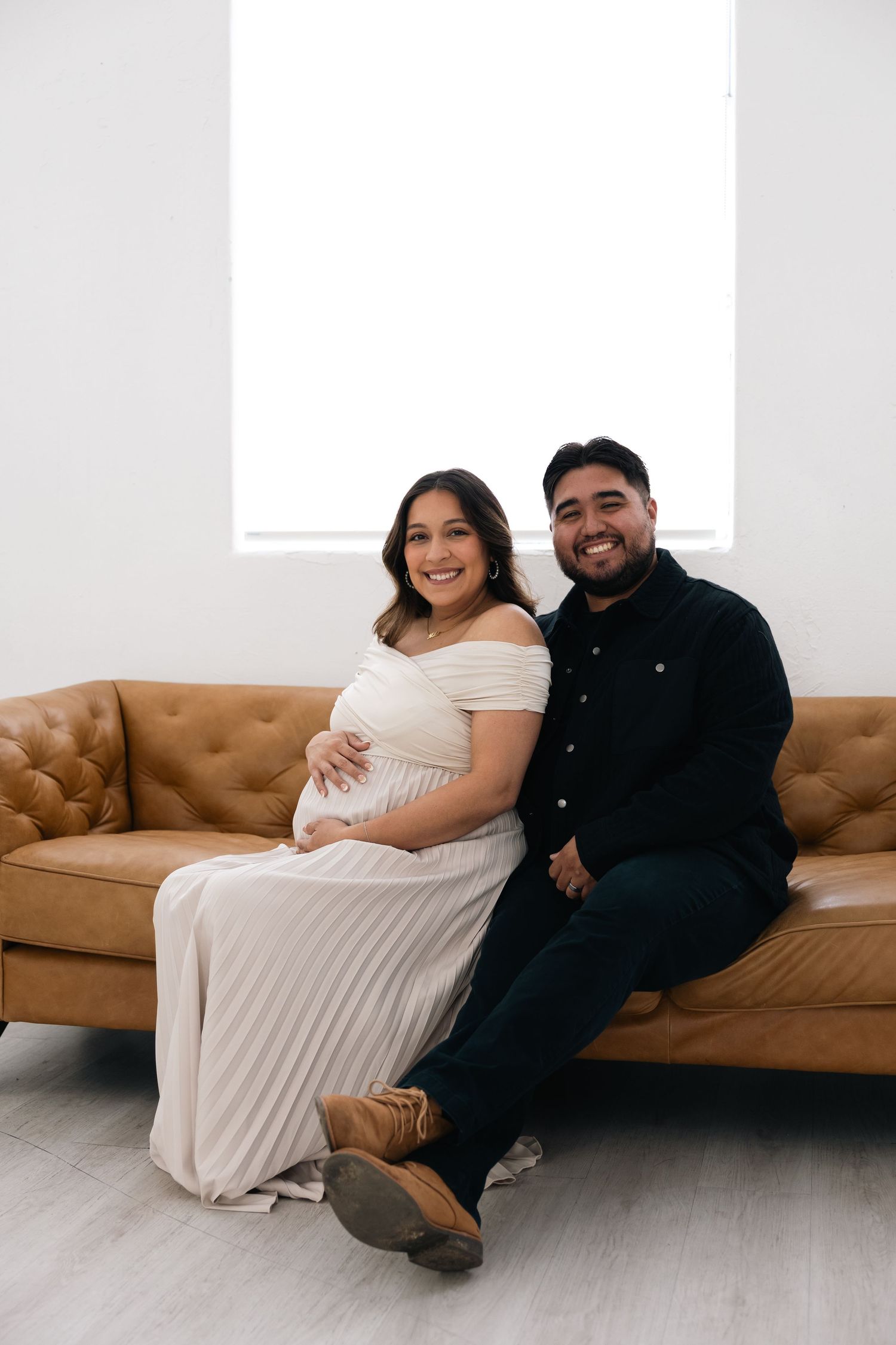 A couple sits together on a brown leather couch in a minimalist indoor setting wearing black and white outfits.